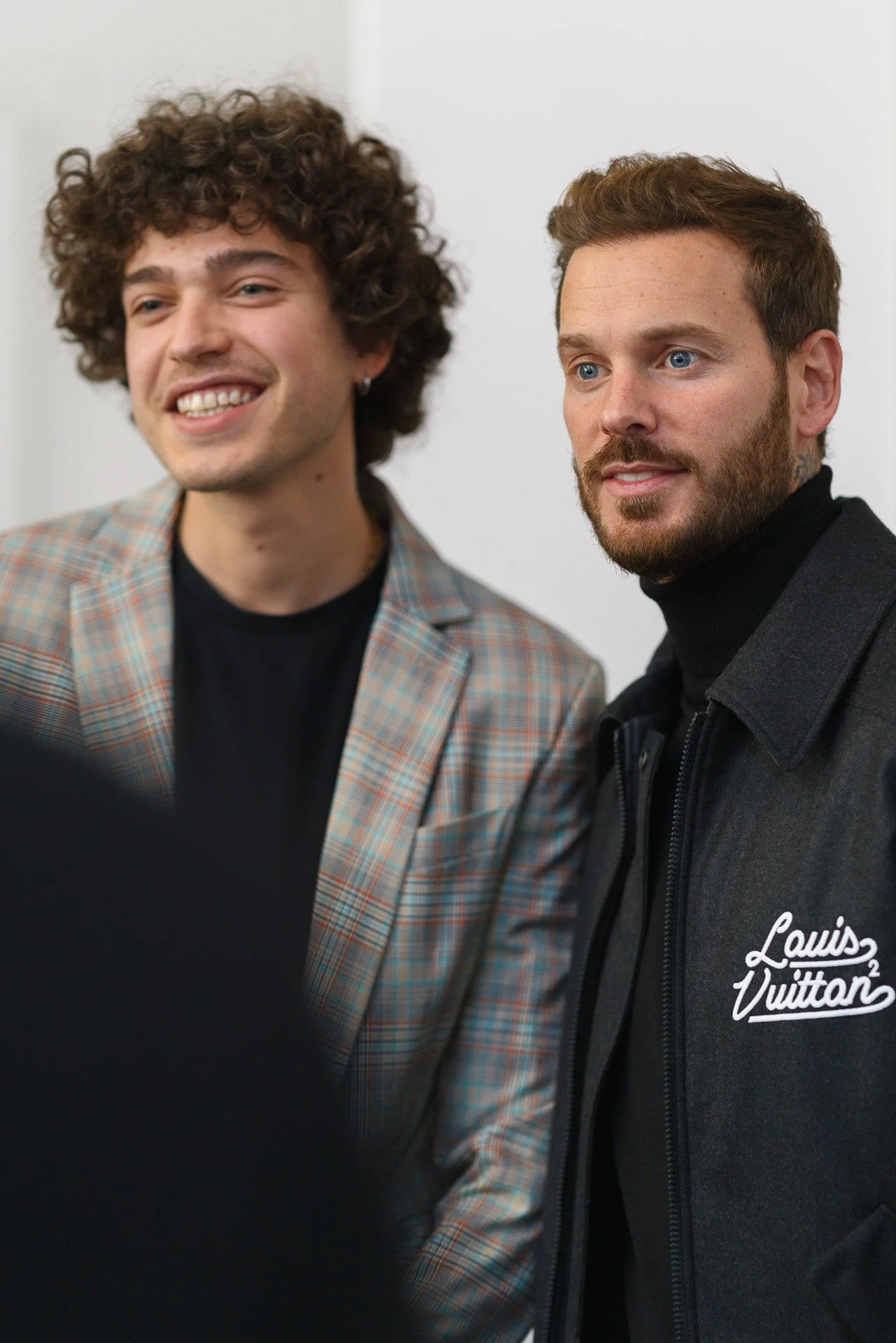 Un homme avec des cheveux bruns et une barbe, portant une veste noire avec le logo Louis Vuitton, tient un micro lors d'une présentation, à côté d'une affiche montrant un produit Braun KS-100.
