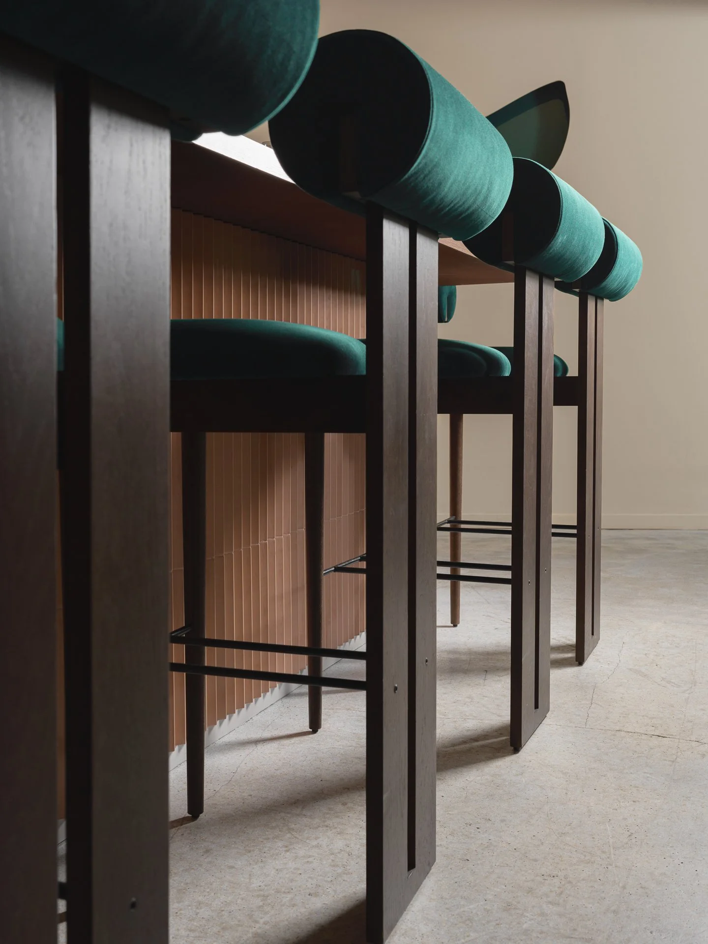 Vue sous un angle bas de trois chaises avec assises vert émeraude, pieds en bois foncé, placées sous un comptoir en bois clair, dans une pièce aux murs beige claire et sol en béton.