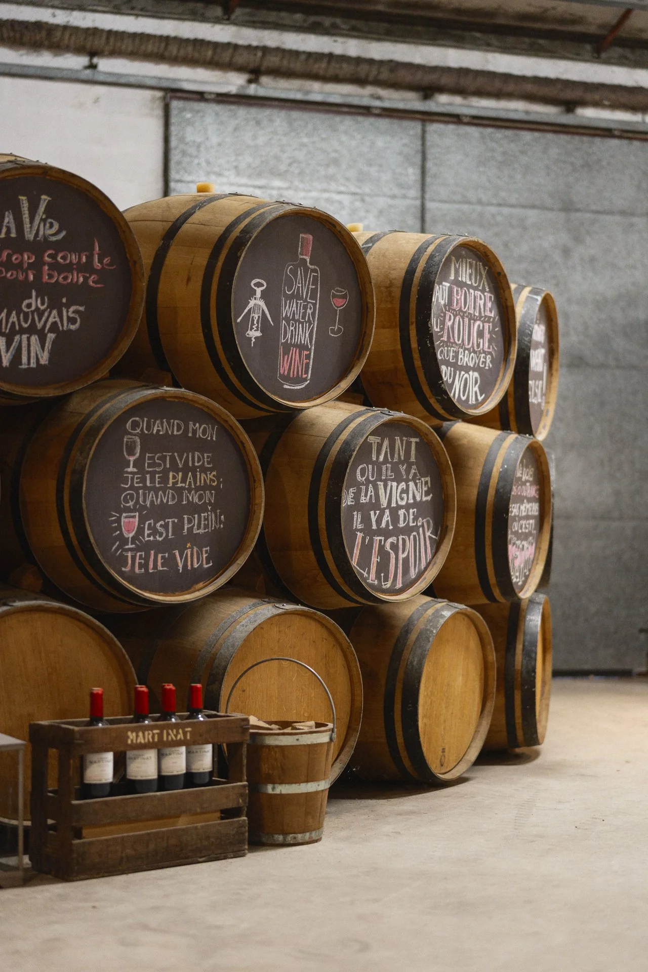 Petits tonneaux de vin avec des messages écrits à la craie sur leurs faces, disposés en rangée dans une cave, avec une caisse de bouteilles de vin en avant.