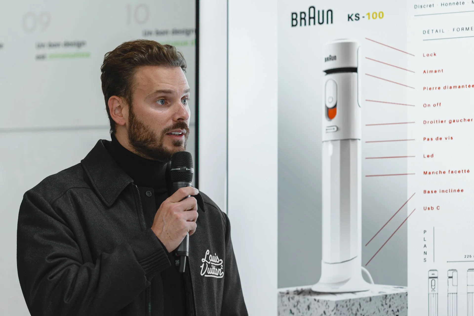 Un homme avec des cheveux bruns et une barbe, portant une veste noire avec le logo Louis Vuitton, tient un micro lors d'une présentation, à côté d'une affiche montrant un produit Braun KS-100.