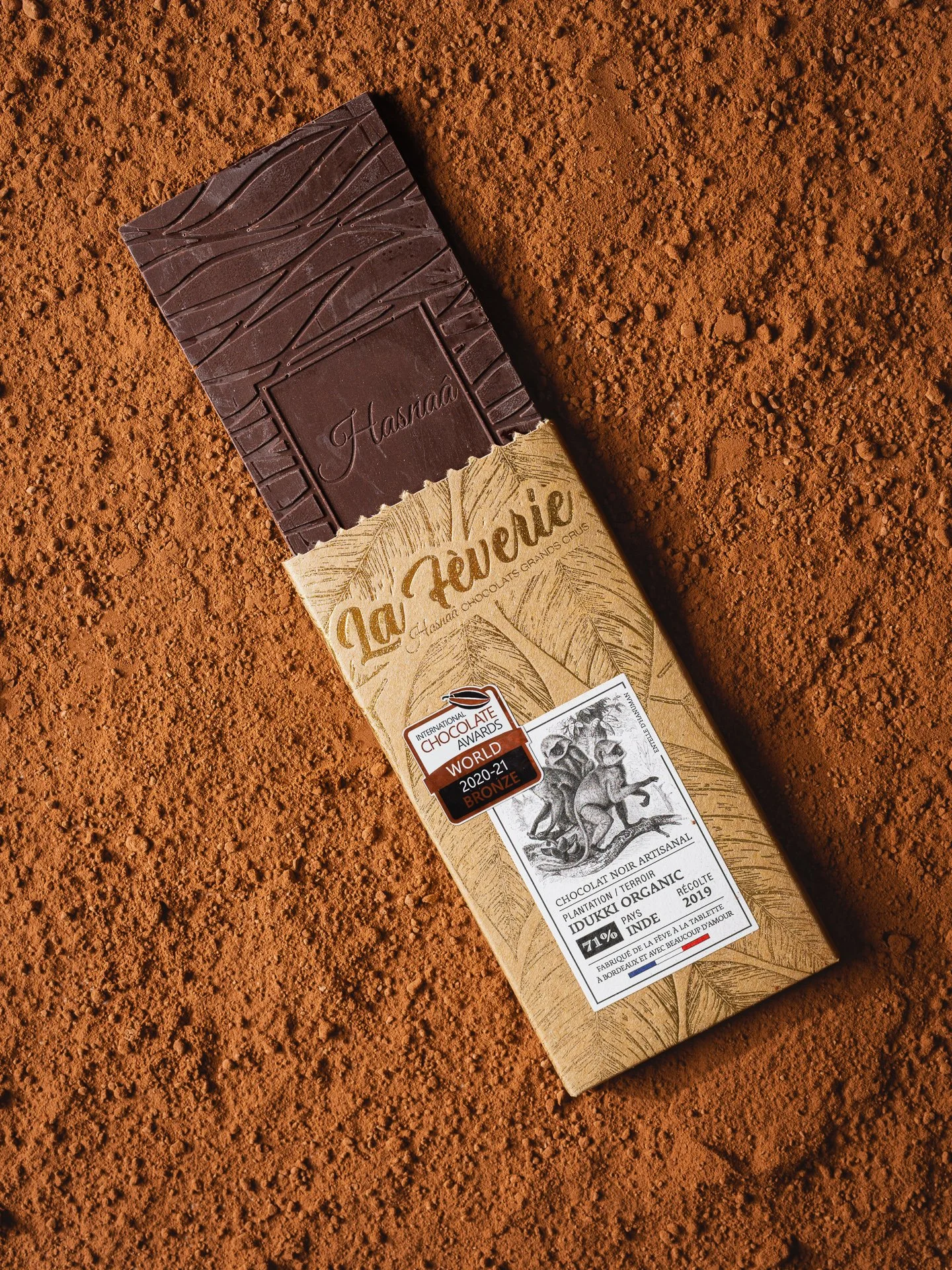 Une tablette de chocolat noir artisanal, emballée dans un emballage beige avec des illustrations de singes et des inscriptions en français. La tablette est posée sur un fond de terre rouge.