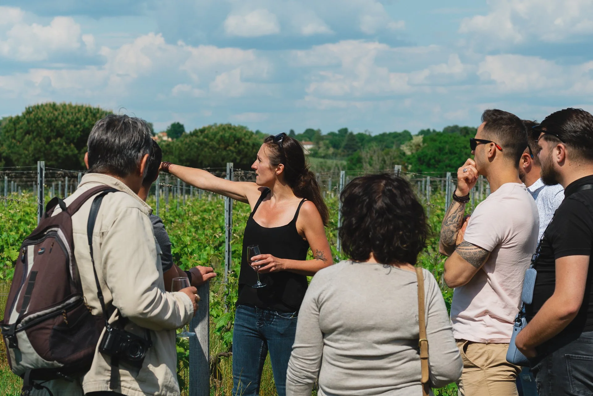 Un groupe de personnes écoutant une femme qui explique la viticulture dans un vignoble en plein air sous un ciel partiellement nuageux.