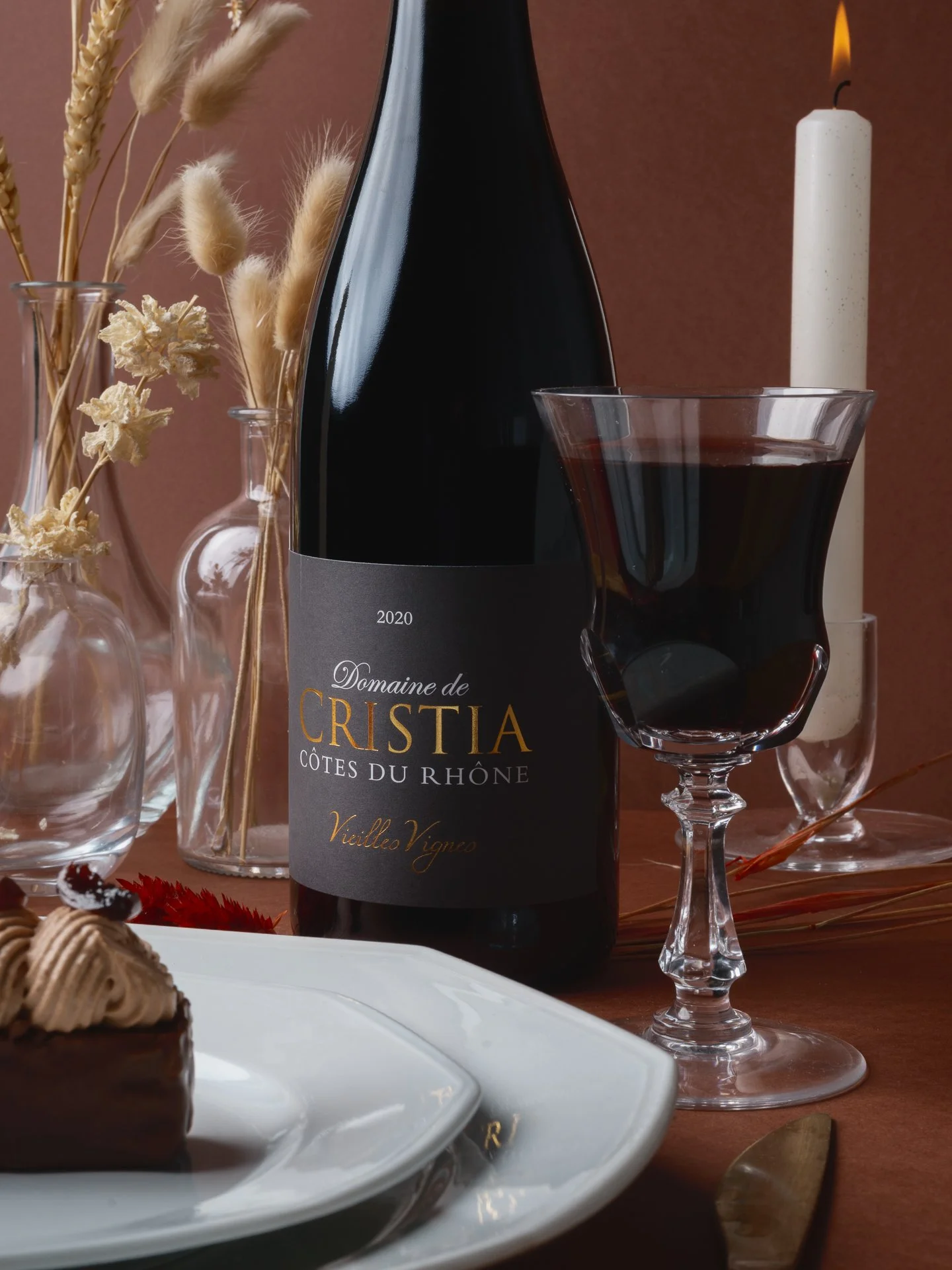 Une bouteille de vin rouge Domaine de Cristia, Côtes du Rhône, Vieilles Vignes, un verre de vin rouge, des vases avec des herbes séchées, une bougie allumée, un gâteau au chocolat avec de la crème et des décors festifs.