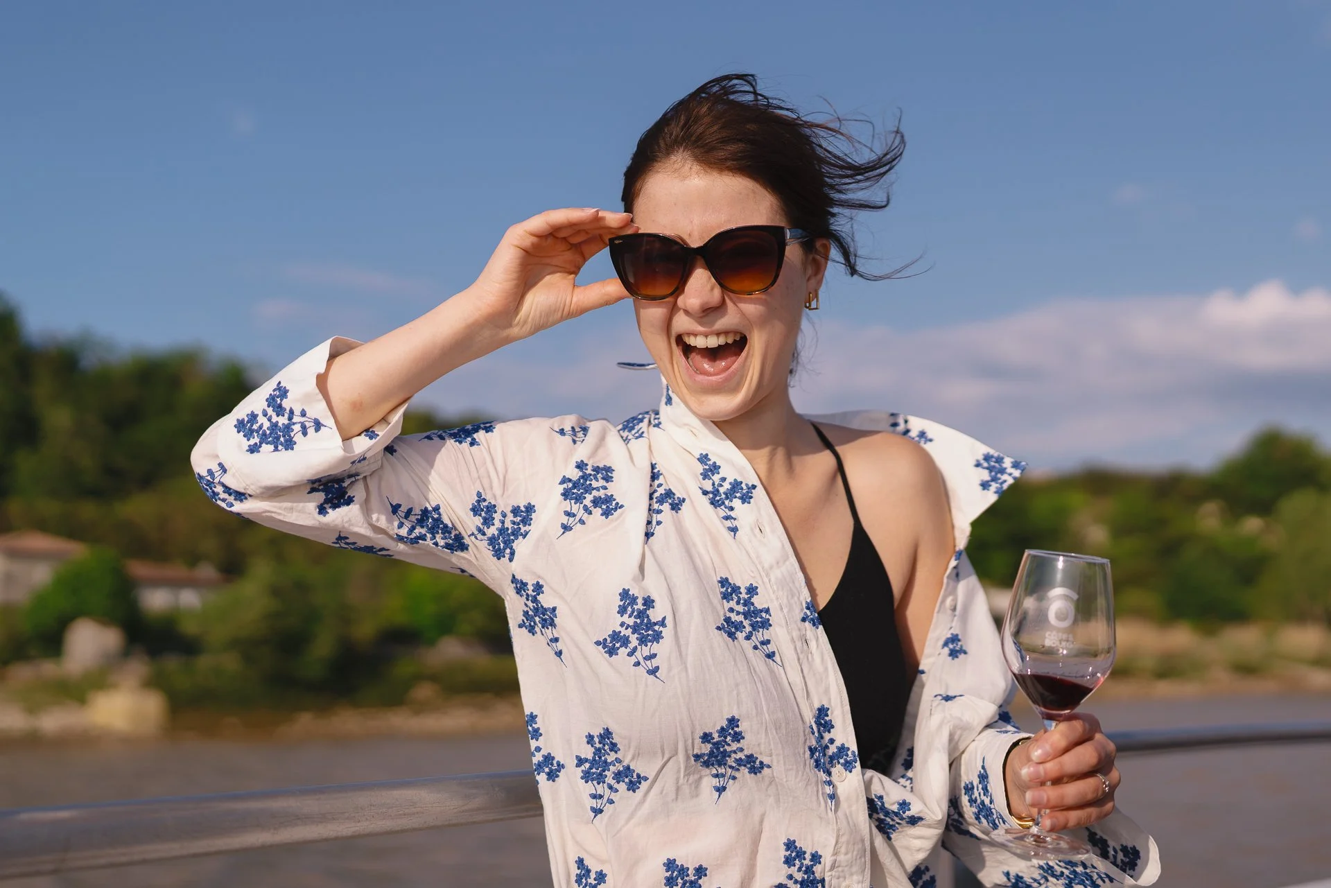 Femme souriante portant des lunettes de soleil, tenant un verre de vin rouge, au bord d'une rivière en plein air, sous un ciel bleu avec quelques nuages.
