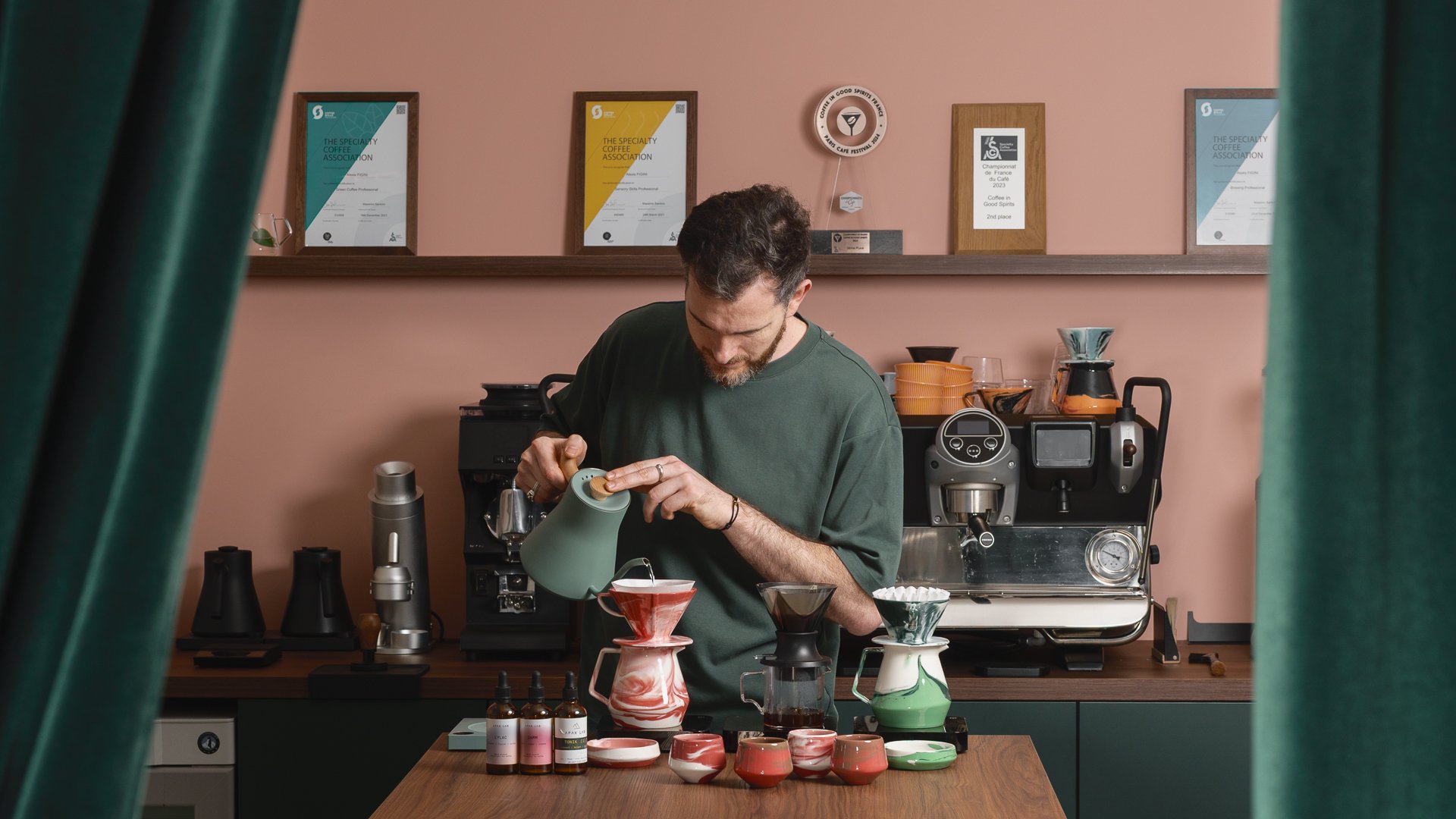 Un homme prépare du café dans un café avec une machine à espresso et diverses tasses colorées sur une table en bois, avec des prix et certificats accrochés au mur en arrière-plan.
