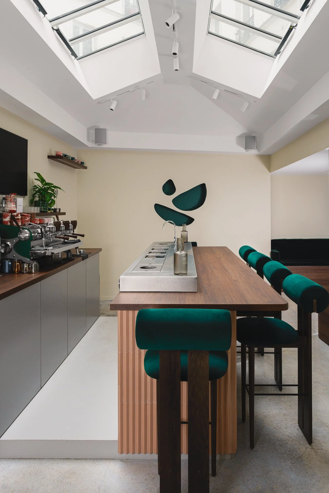 Intérieur moderne d'une cuisine ou un bar avec un plafond en pente avec des fenêtres de toit. Il y a un comptoir en bois avec des tabourets en velours vert foncé. Le mur en arrière-plan a une décoration abstraite en forme de morceaux de verre ou d'ar
