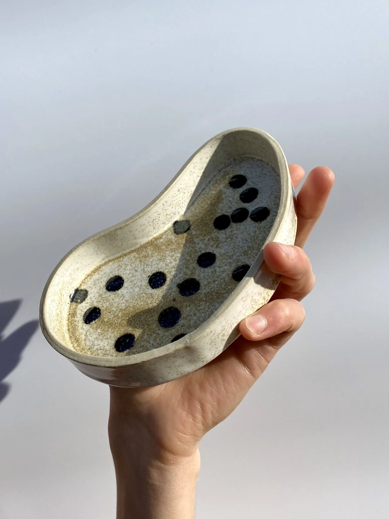 A hand holds a ceramic kidney-shaped dish with off-white glaze and metallic black spots in front of a white backdrop.