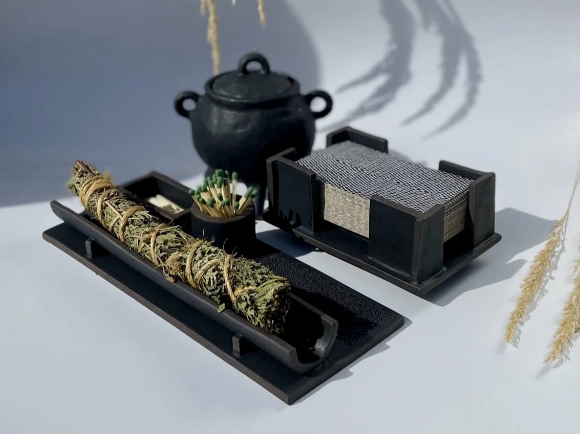 A rectangular ceramic tray holding a bundle of dried herbs with a match jar and striker, next to a ceramic tarot deck holder and a black ceramic cauldron on a white backdrop.