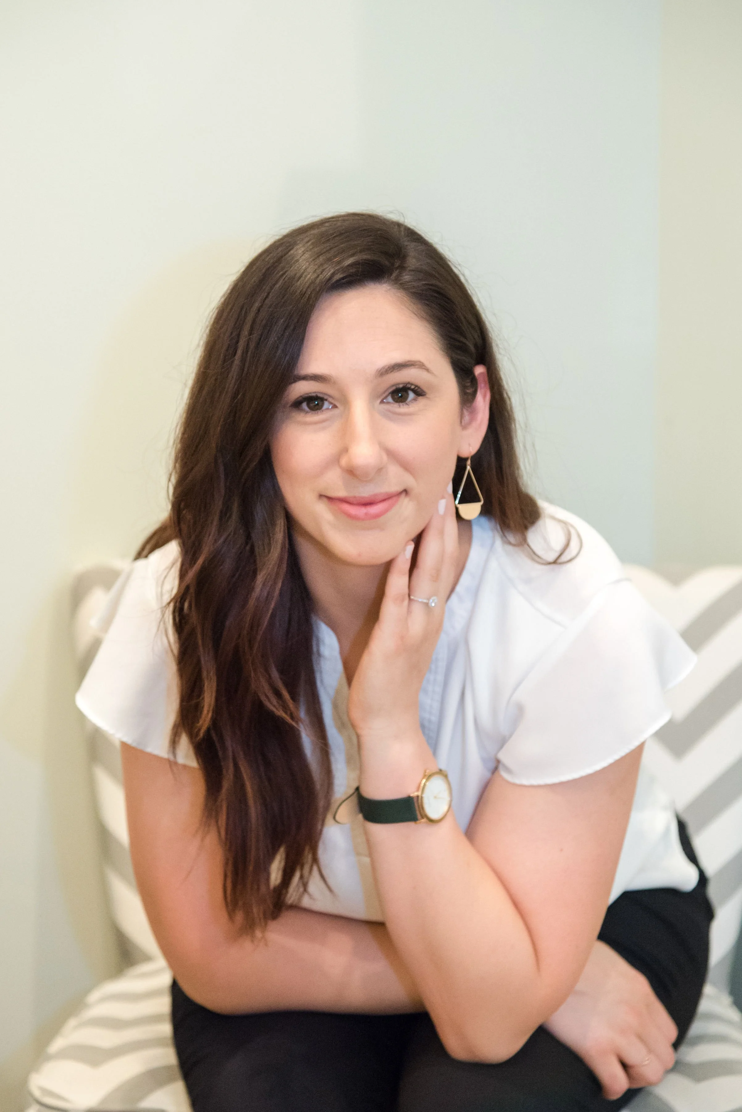 A young woman with long, dark brown hair, wearing a white top, black shorts, and gold jewelry, sitting on a bed with a striped blanket, smiling and resting her chin on her hand.