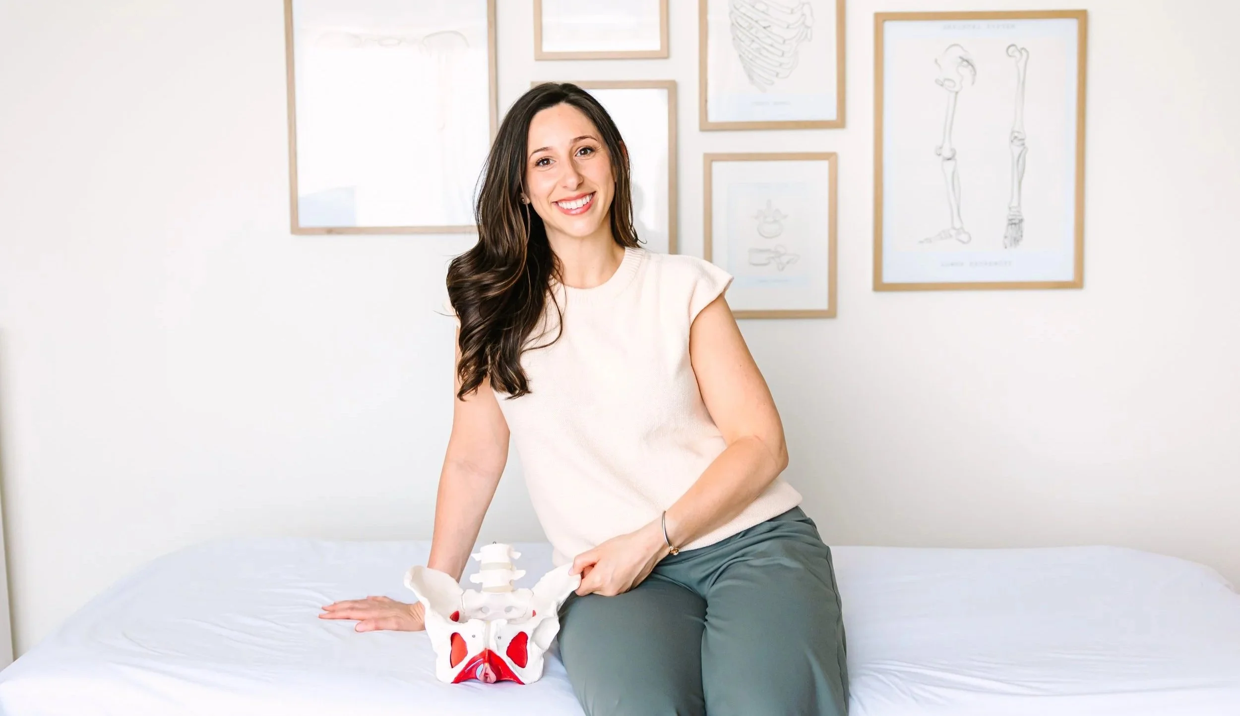 A smiling woman with long dark hair sits on a bed in a room with framed anatomical sketches on the wall. She has her left hand on a model of a pelvis and spine.