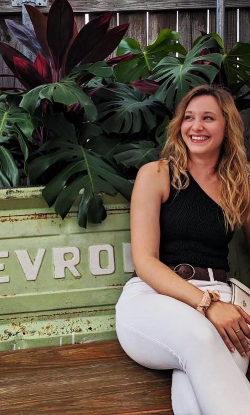 A woman with curly blonde hair smiling and sitting outdoors on a wooden bench in front of lush green and purple tropical plants. She is wearing a sleeveless green top and white pants.