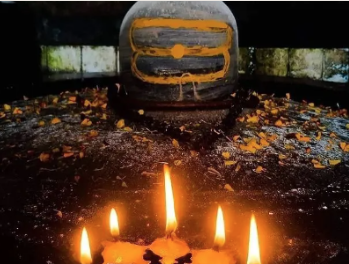 A burning candle surrounded by small flames with a large train wheel in the background, located in a dark tunnel with some light coming through the sides.
