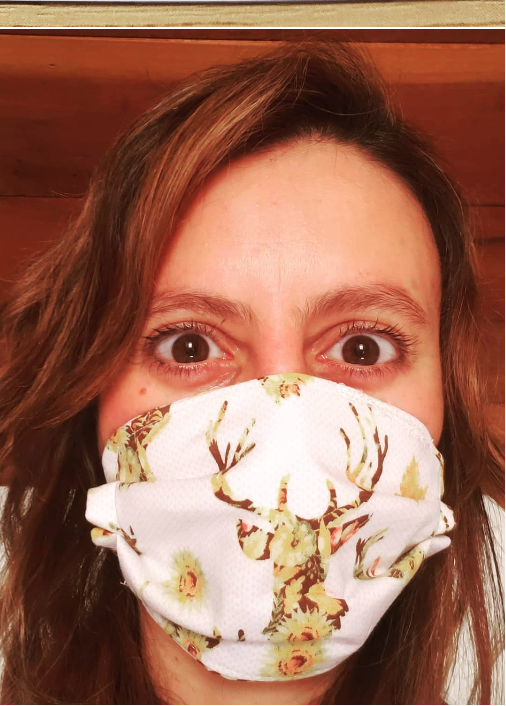 A woman with brown hair takes a selfie wearing a floral face mask with a deer design, showing her wide eyes and eyebrows.