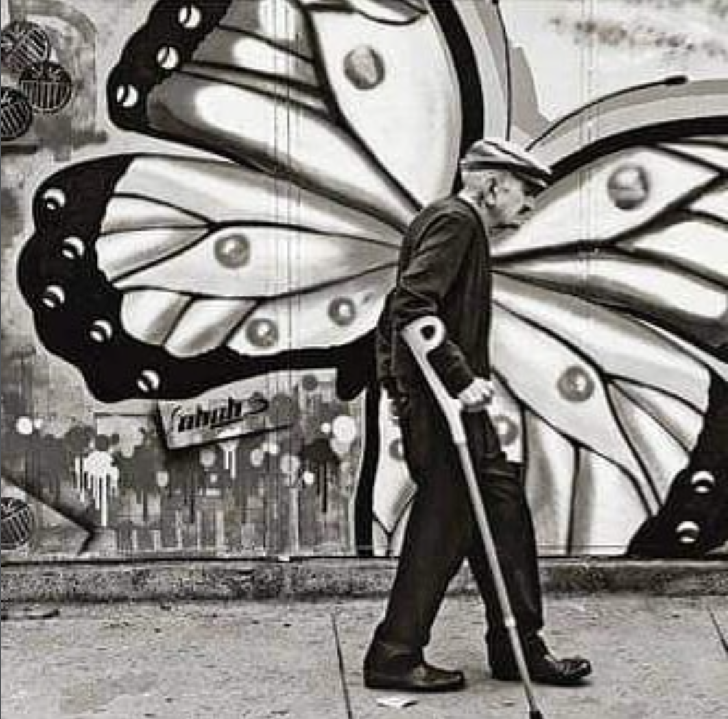 A man wearing a hat, jacket, and glasses, walking with a crutch, in front of a mural of a large butterfly with detailed wing patterns painted in black and white on a wall.