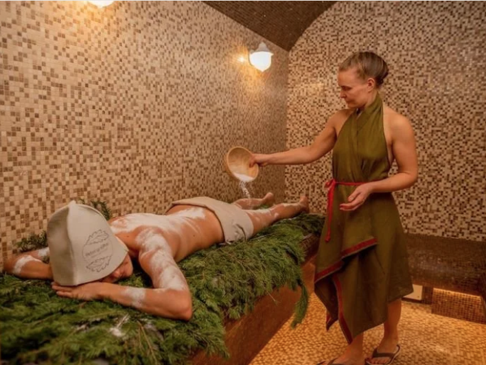 A woman giving a salt scrub massage to a man lying face down on a treatment table in a spa room with warm lighting and tiled walls.