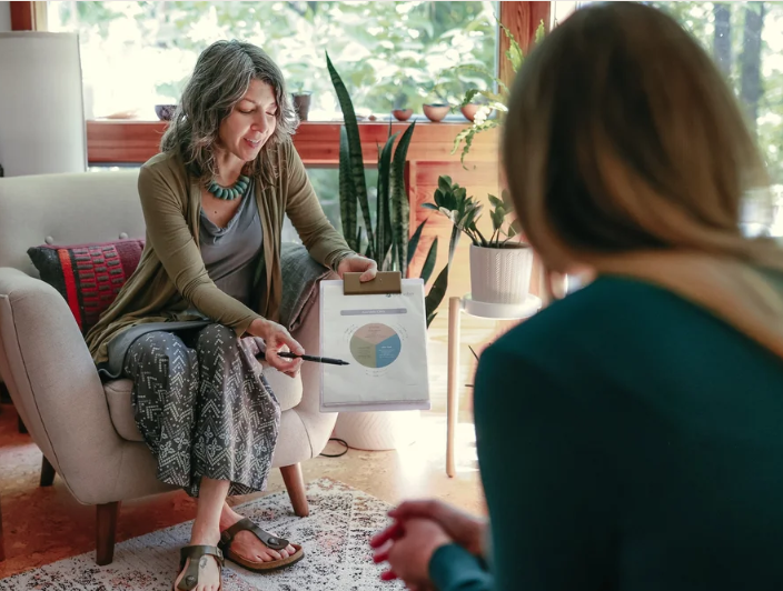 Two women having a conversation in a cozy, well-lit living room. One woman is sitting on a sofa, holding a pen and a clipboard with a chart, talking to the other woman who is sitting nearby.