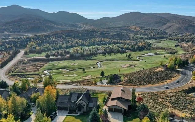 Aerial view of a residential area with houses surrounded by lush greenery and a golf course. Rolling hills and winding roads are visible, with mountains in the background under a clear blue sky.