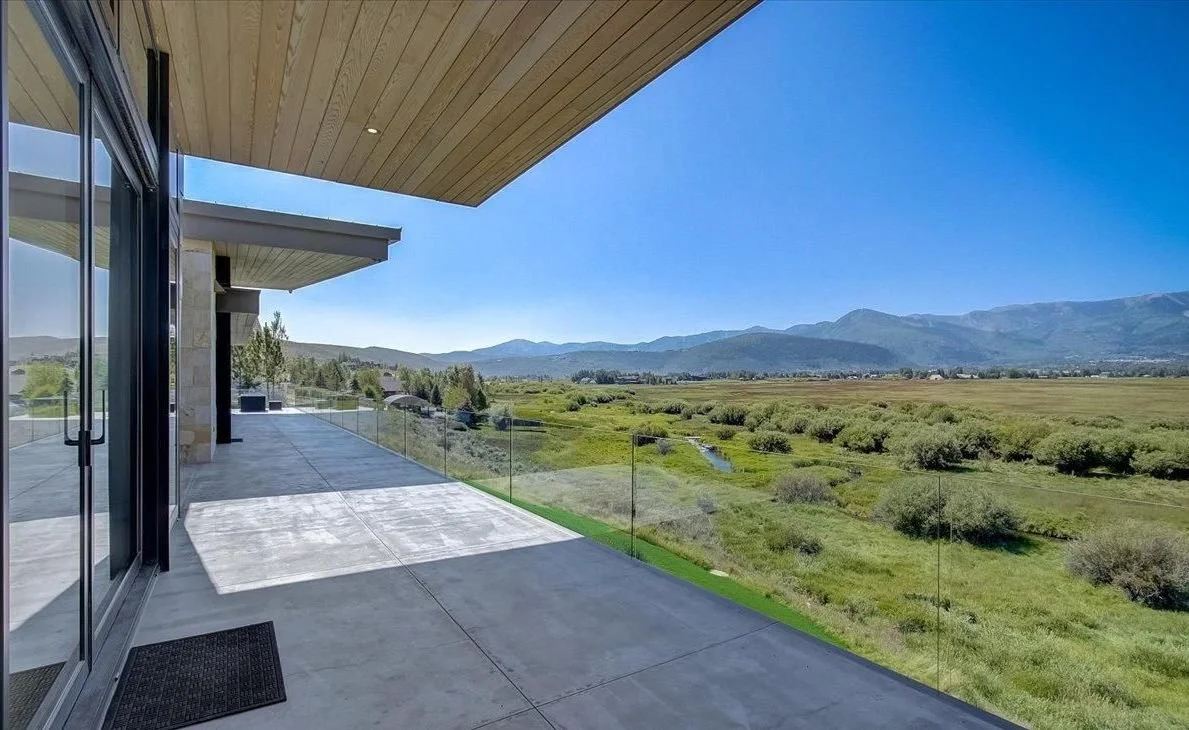 Modern terrace with glass railing overlooking a green landscape and distant mountains under a clear blue sky.