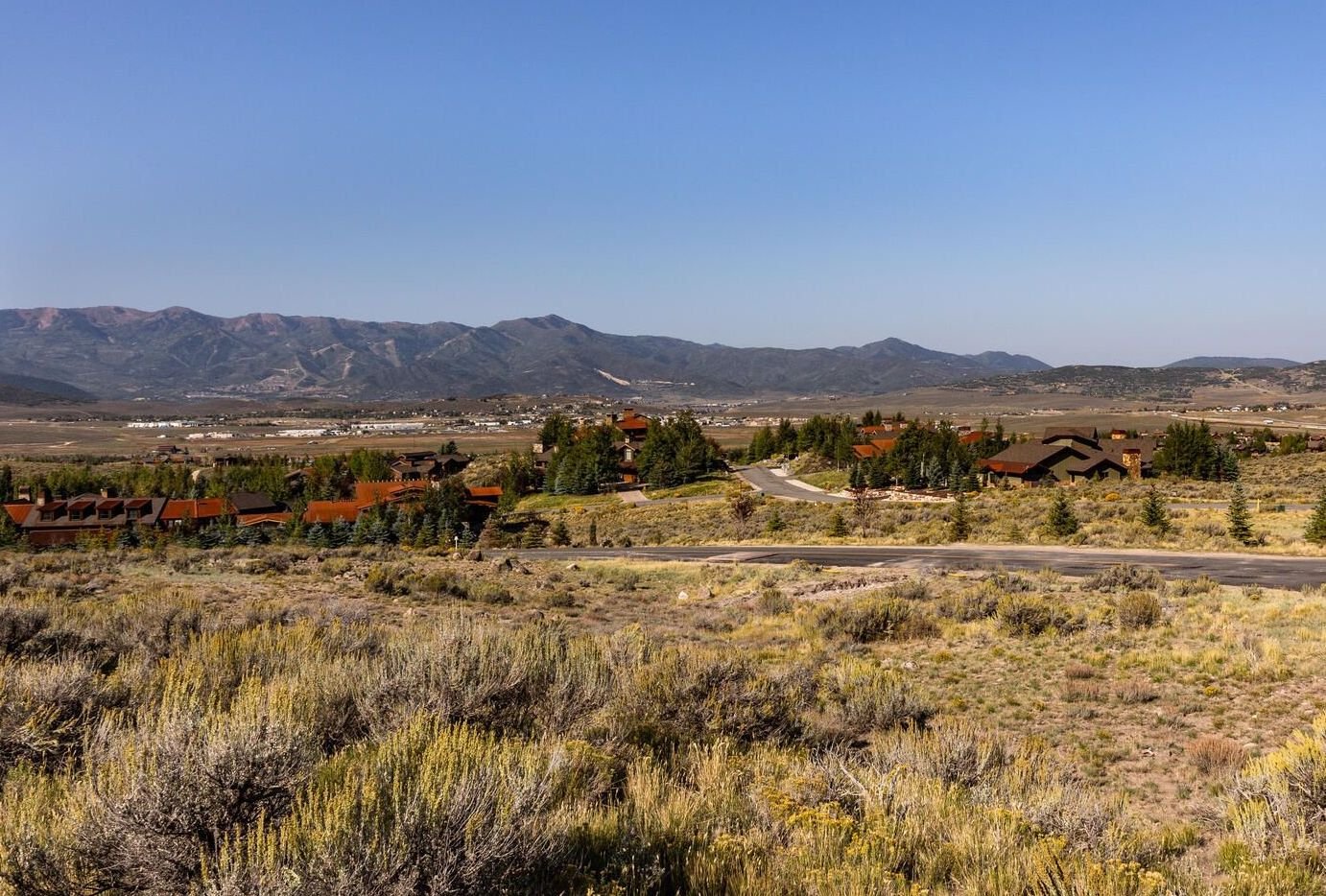 Mountain landscape with houses and shrubs in foreground, road winding through community, distant mountains under clear blue sky.