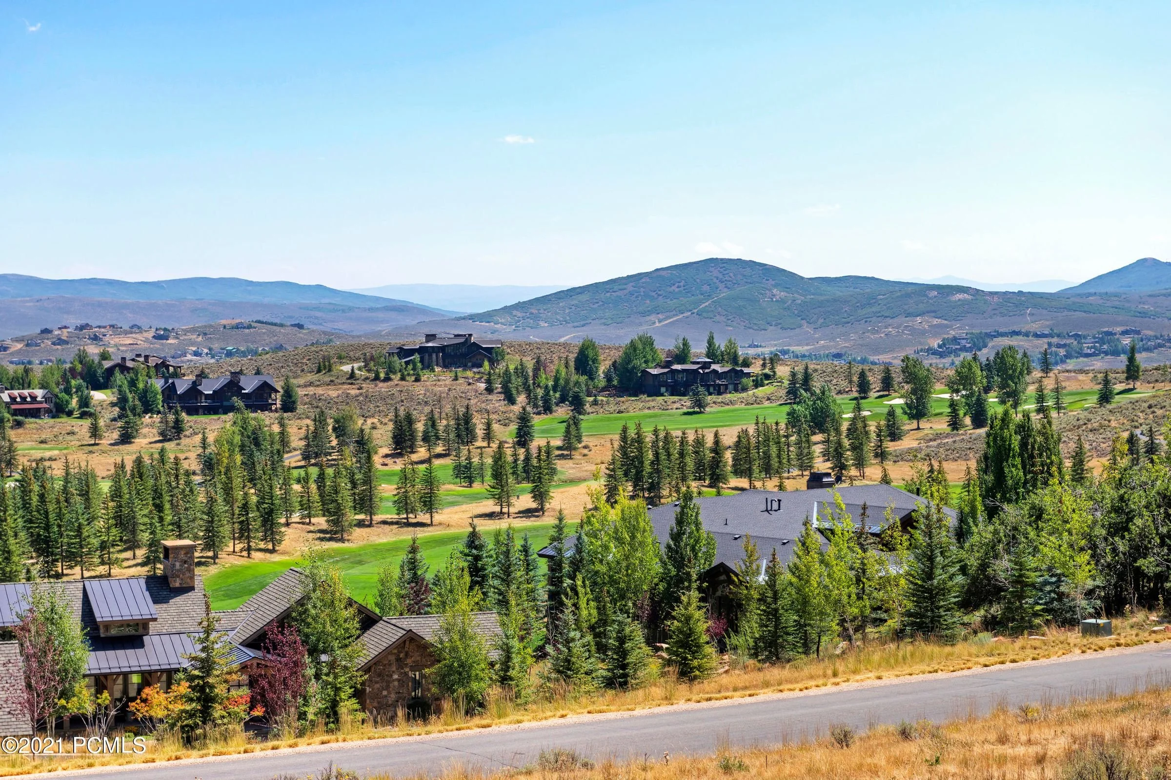 Panoramic view of a residential area with houses, trees, and hills, set against a clear blue sky.
