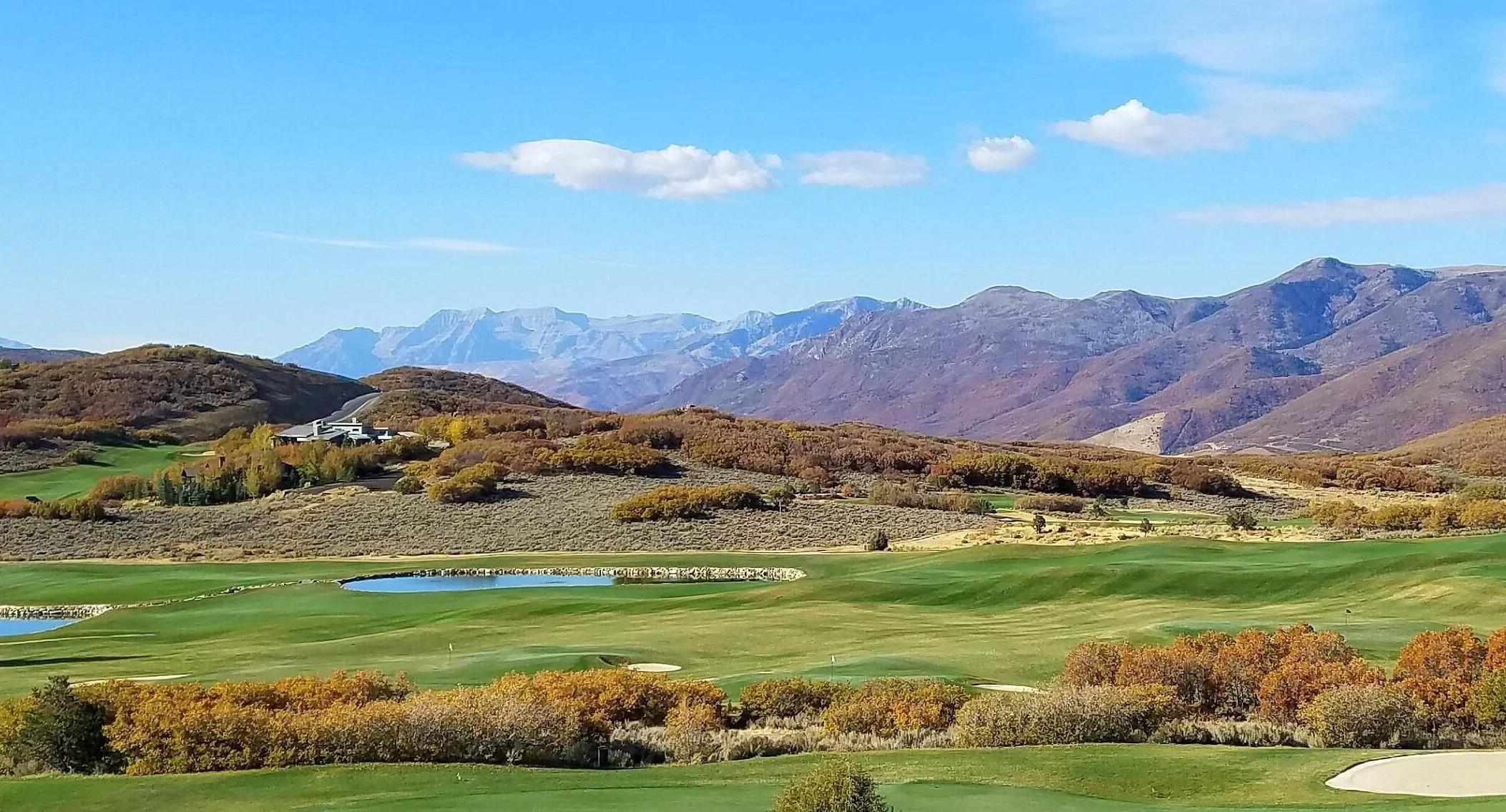 Scenic landscape with a golf course, autumn foliage, and mountain range under a blue sky.