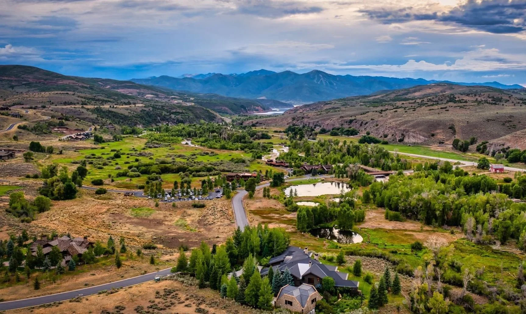 Aerial view of a scenic valley with green fields, trees, and houses, surrounded by hills and mountains in the background. A winding road and several small ponds are visible, under a partly cloudy sky.