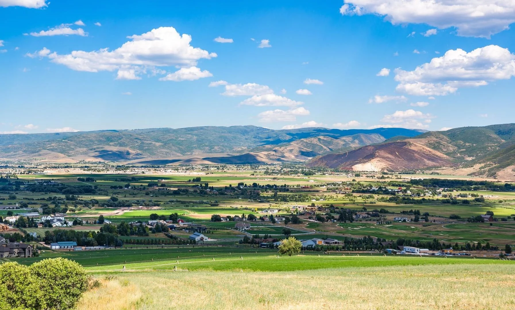 Panoramic view of a verdant valley with farms, fields, and scattered houses. Rolling hills and distant mountains under a blue sky with fluffy clouds.