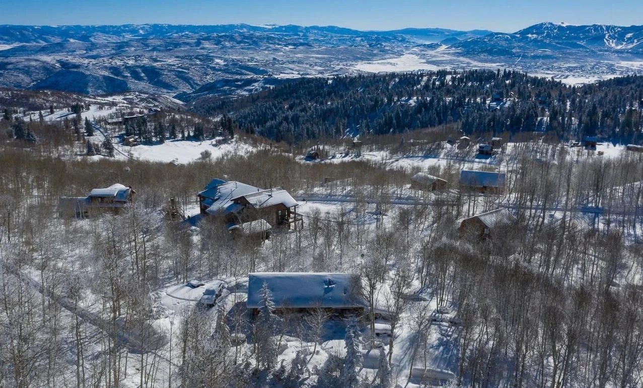 Aerial view of a snowy landscape with cabins and trees, mountains in the background, under a clear blue sky.