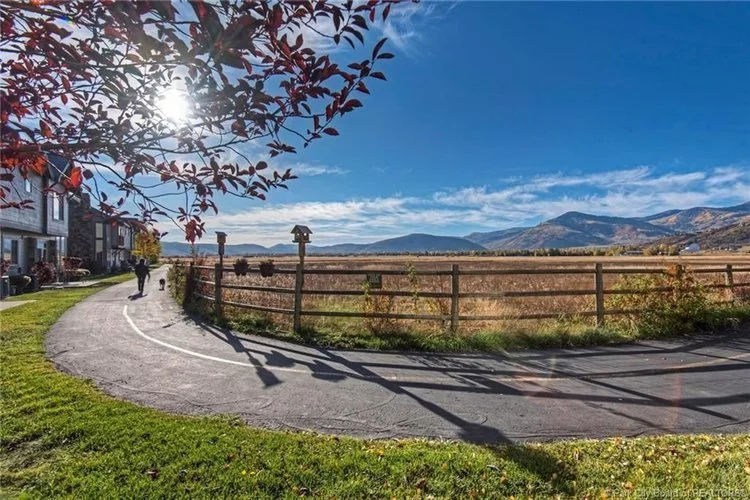 Scenic view of a paved pathway curving through a grassy yard, with a wooden fence and open field stretching toward distant mountains under a clear blue sky, with sunlight filtering through tree branches.