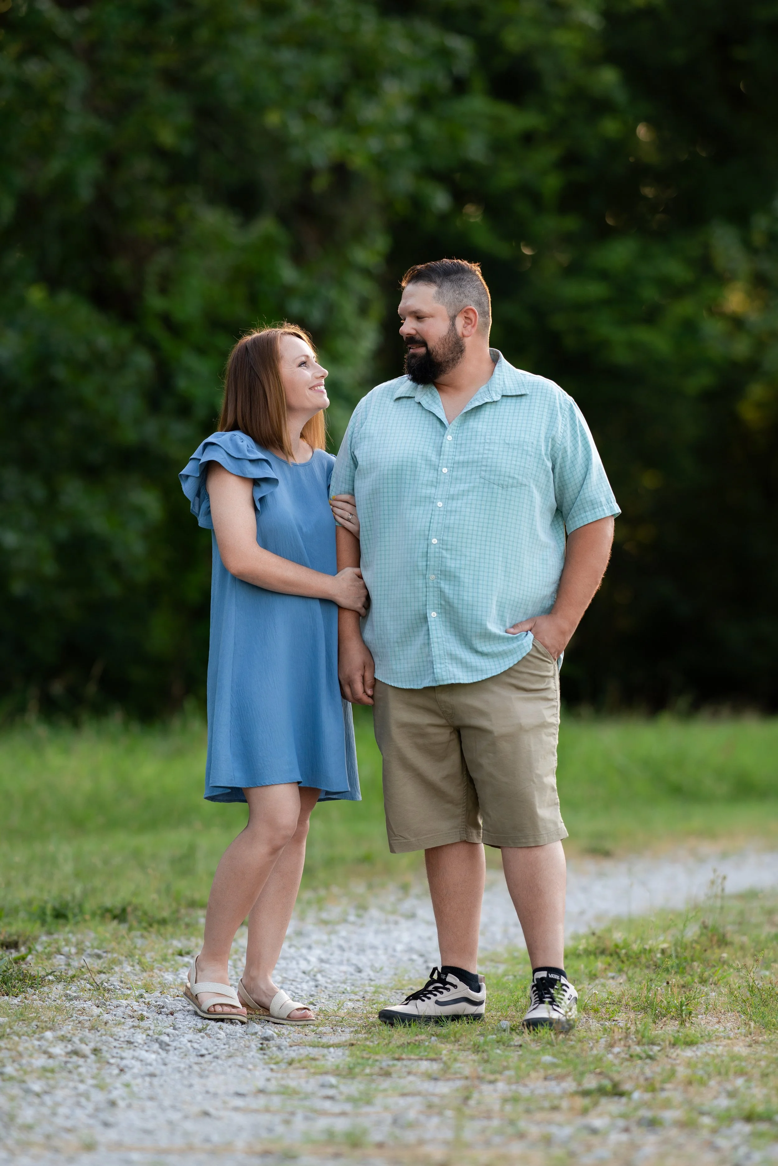 A man and woman standing outdoors on a nature trail, smiling and looking at each other.