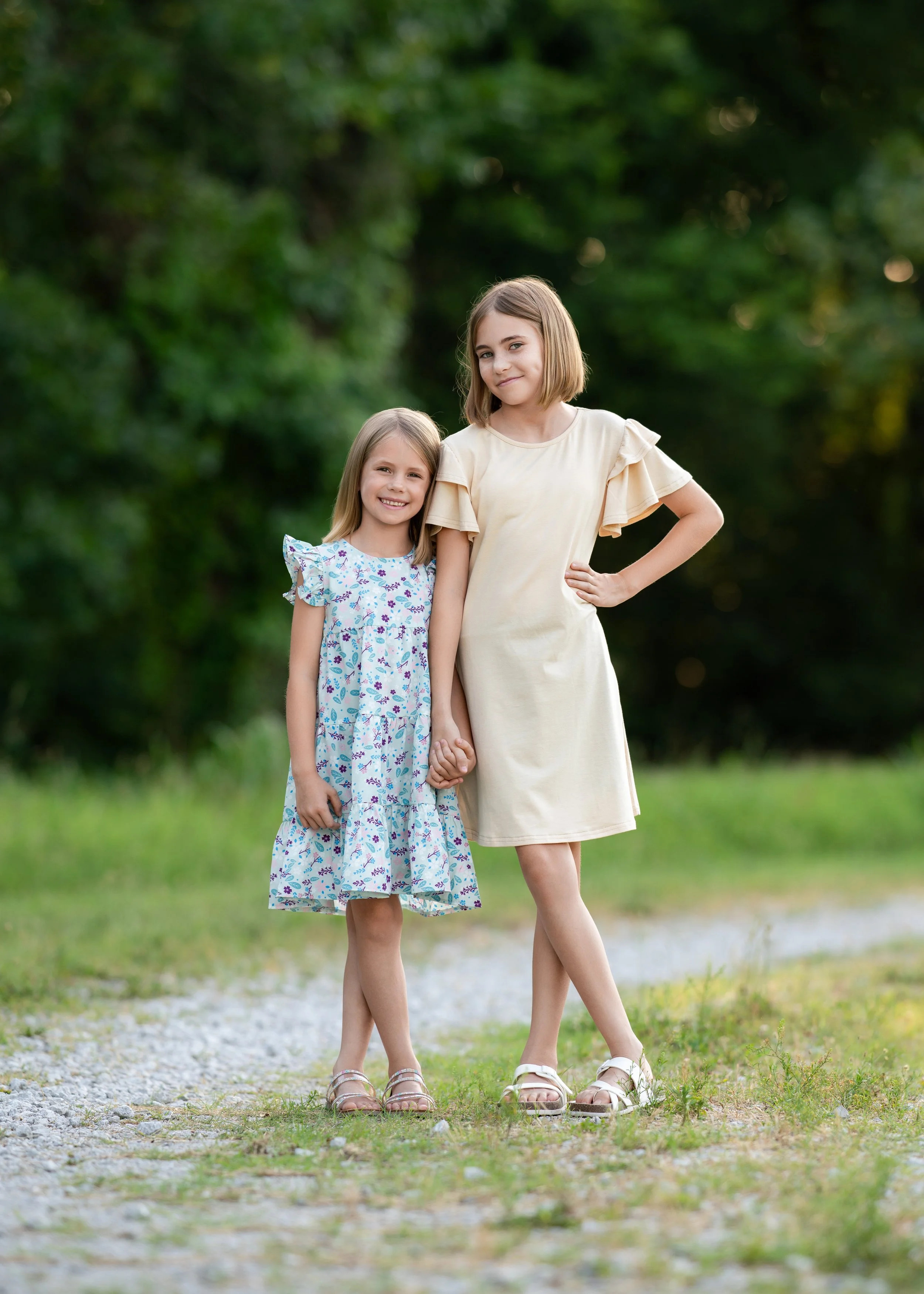 Two young girls standing outdoors on a gravel path, holding hands, with greenery in the background. The older girl is wearing a cream-colored dress with ruffled sleeves, and the younger girl is in a patterned dress with flutter sleeves.