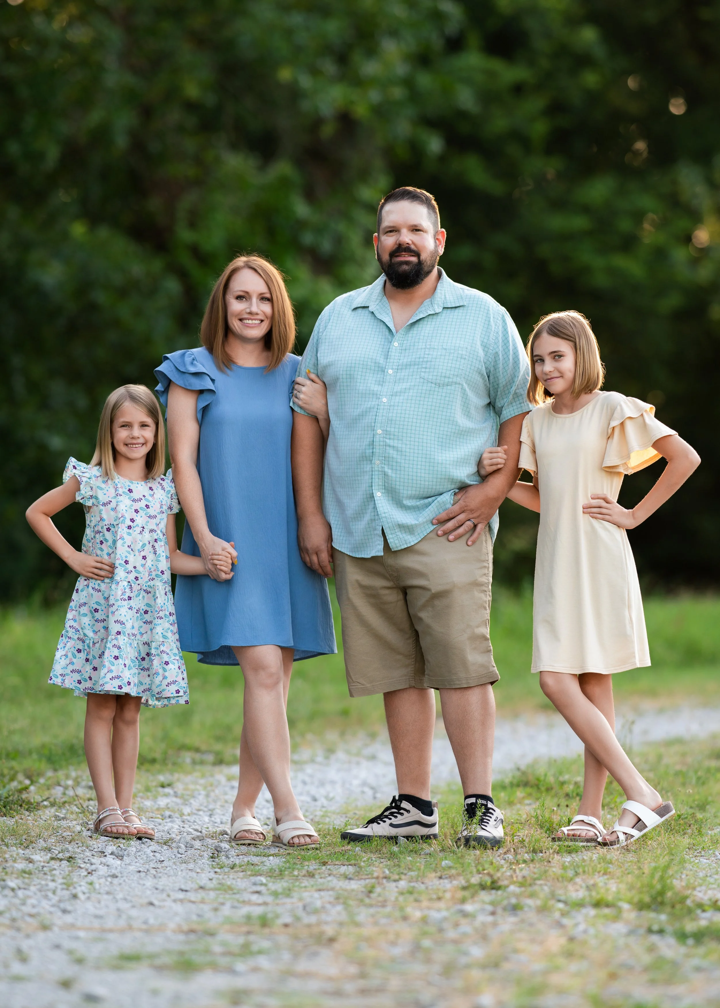 Family of four standing outdoors on a gravel path with green trees in the background. Two young girls and two adults, smiling and holding hands.