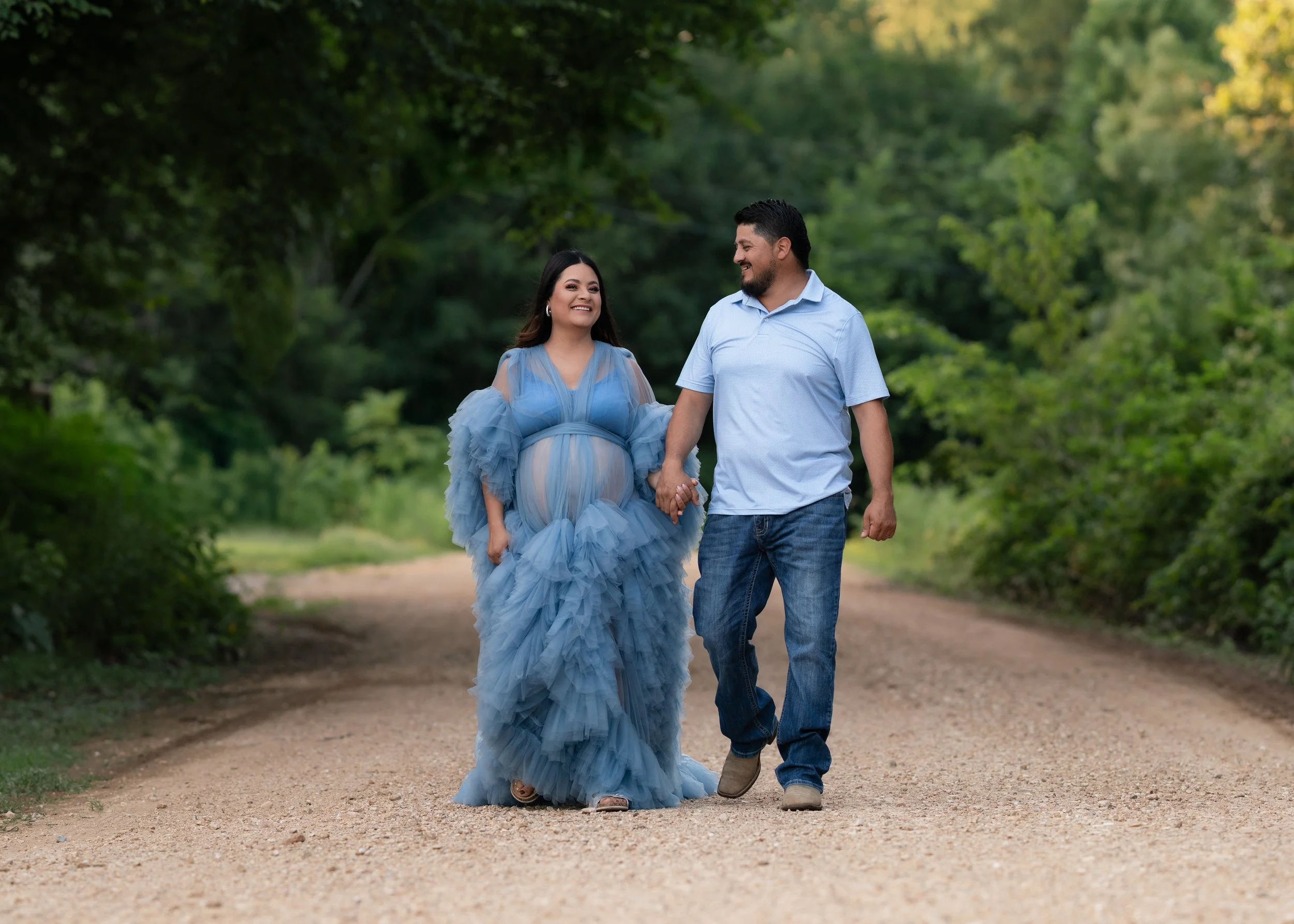 A couple holding hands, walking on a dirt path through a lush green forest, smiling at each other. The woman is wearing a flowing blue maternity gown, and the man is dressed casually in a light blue shirt and jeans.