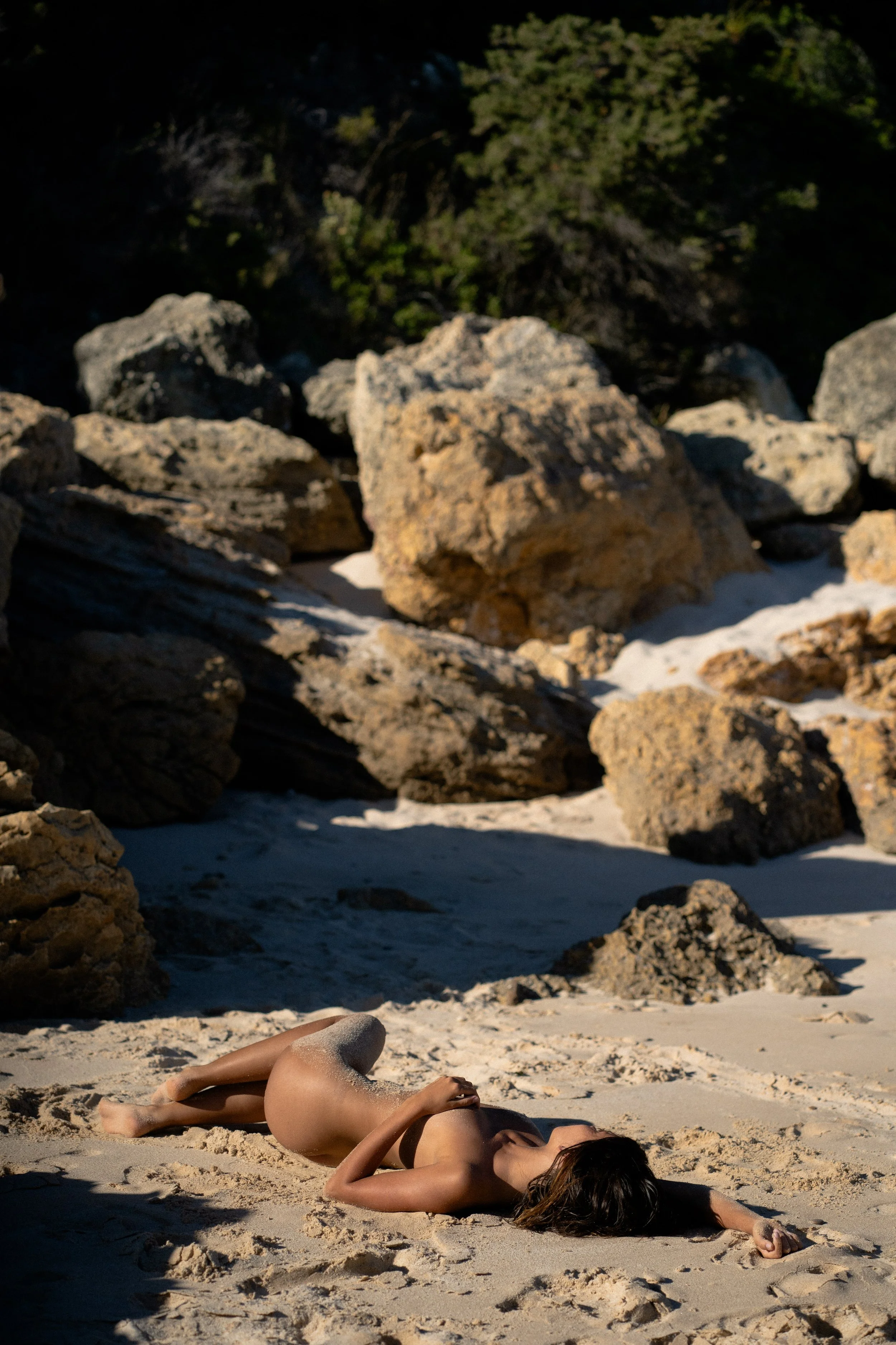 A woman lying on the sandy beach with her head resting on her arm, surrounded by rocks and trees in the background.