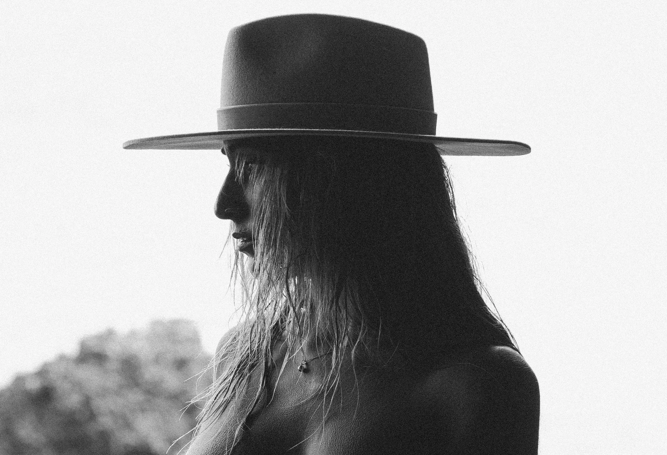 Black and white profile of a woman wearing a wide-brimmed hat and a necklace, with a blurred natural landscape background.