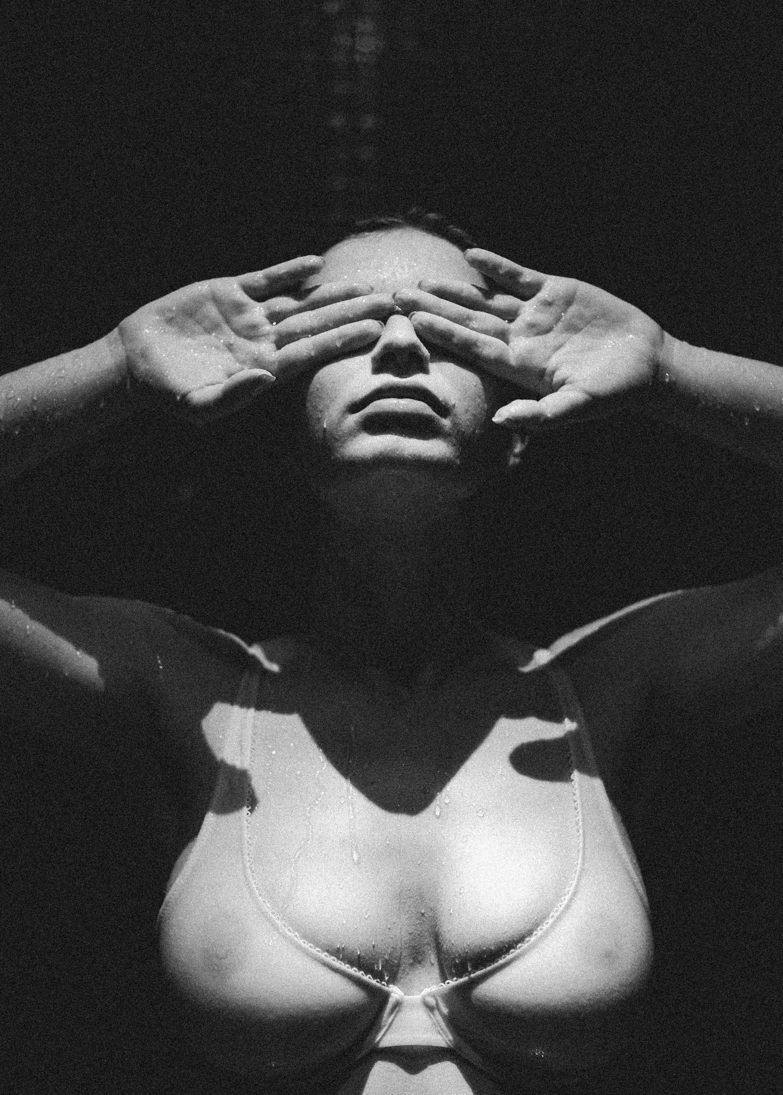 A black and white photo of a woman with wet hair and water droplets on her face and chest, covering her eyes with her hands.