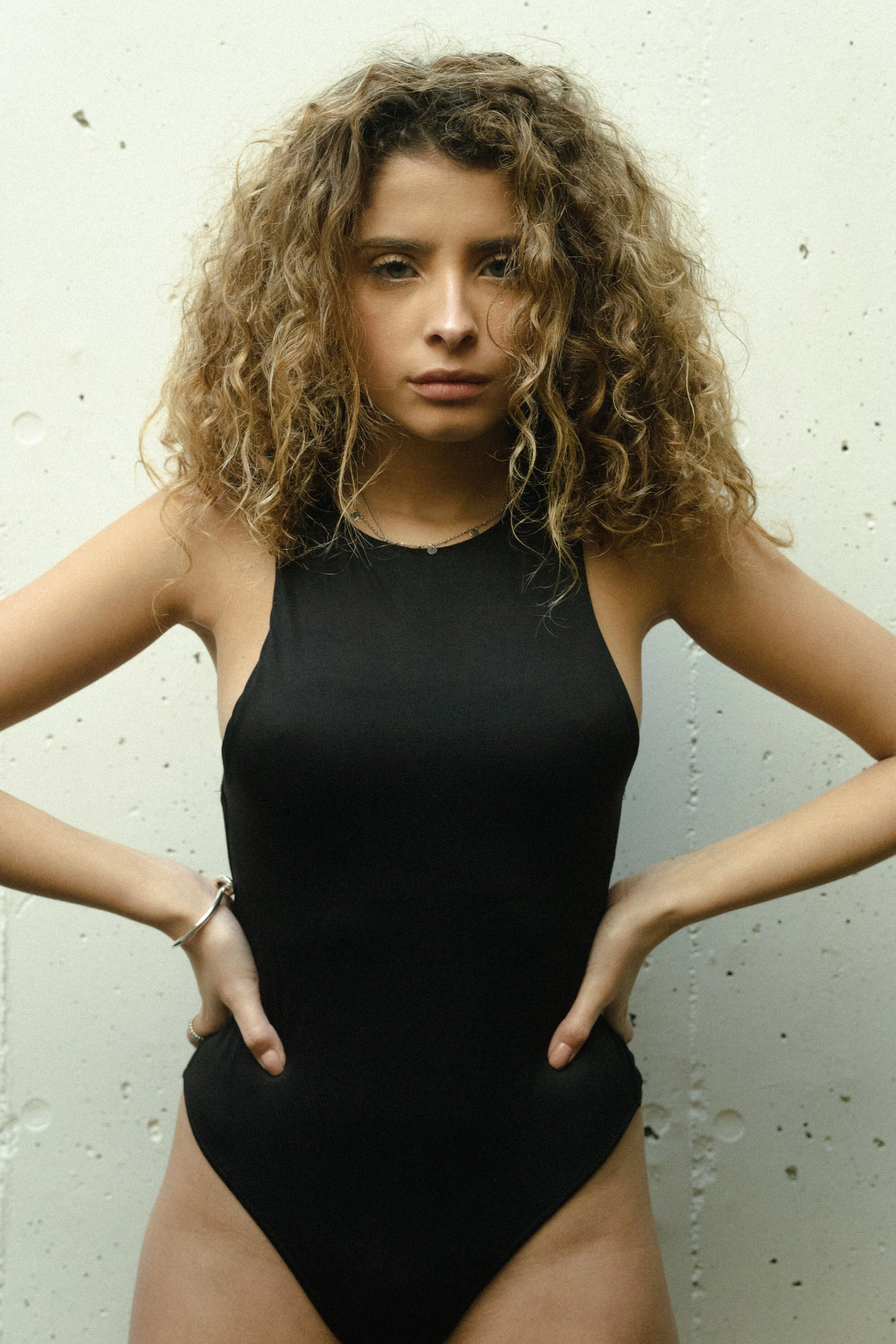 A woman with curly hair wearing a black sleeveless top and jewelry, standing against a white wall with small black marks, with her hands on her hips.