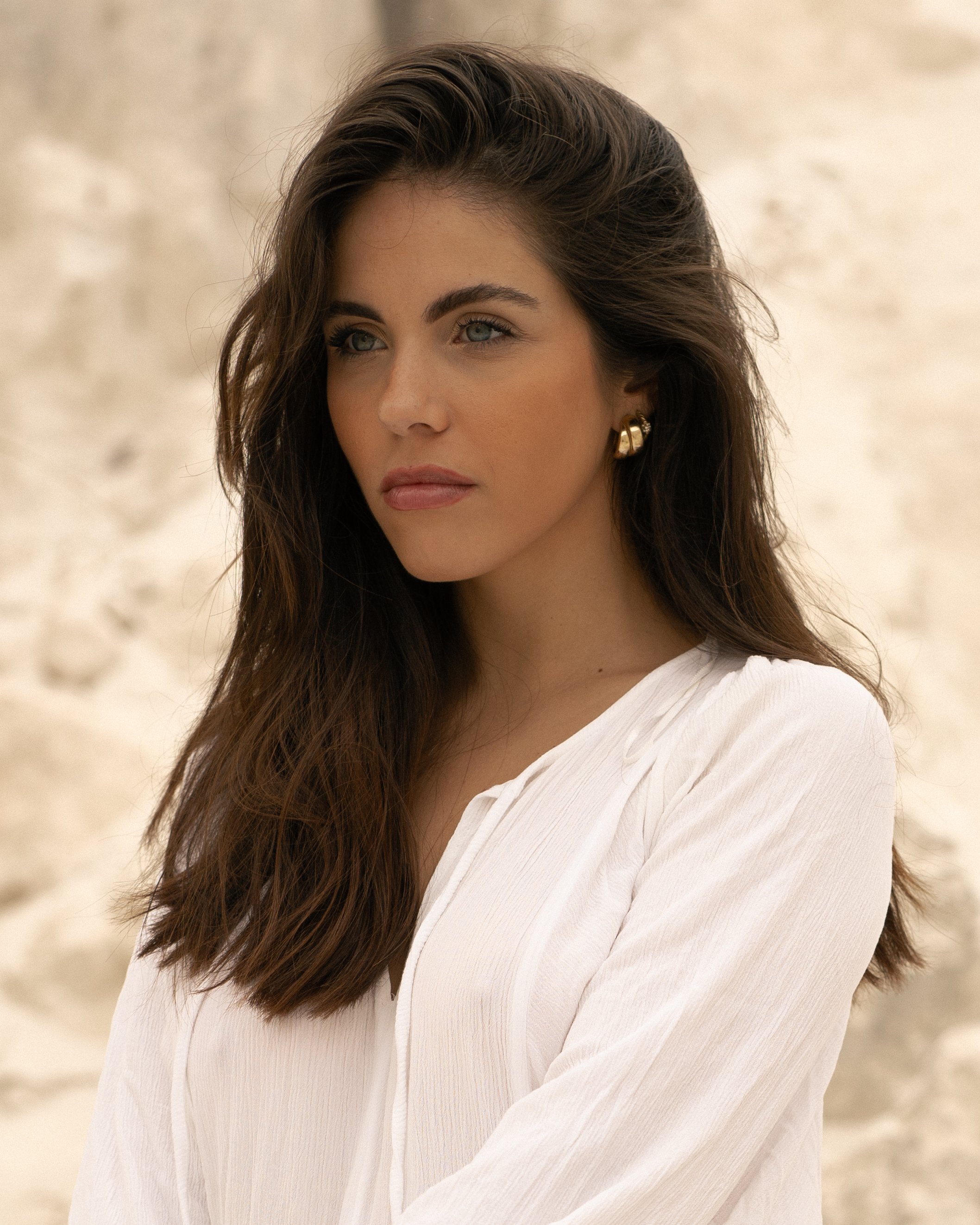 A woman with long brown hair, light eye makeup, and gold earrings, wearing a white textured top, looking to her left, standing against a beige textured background.