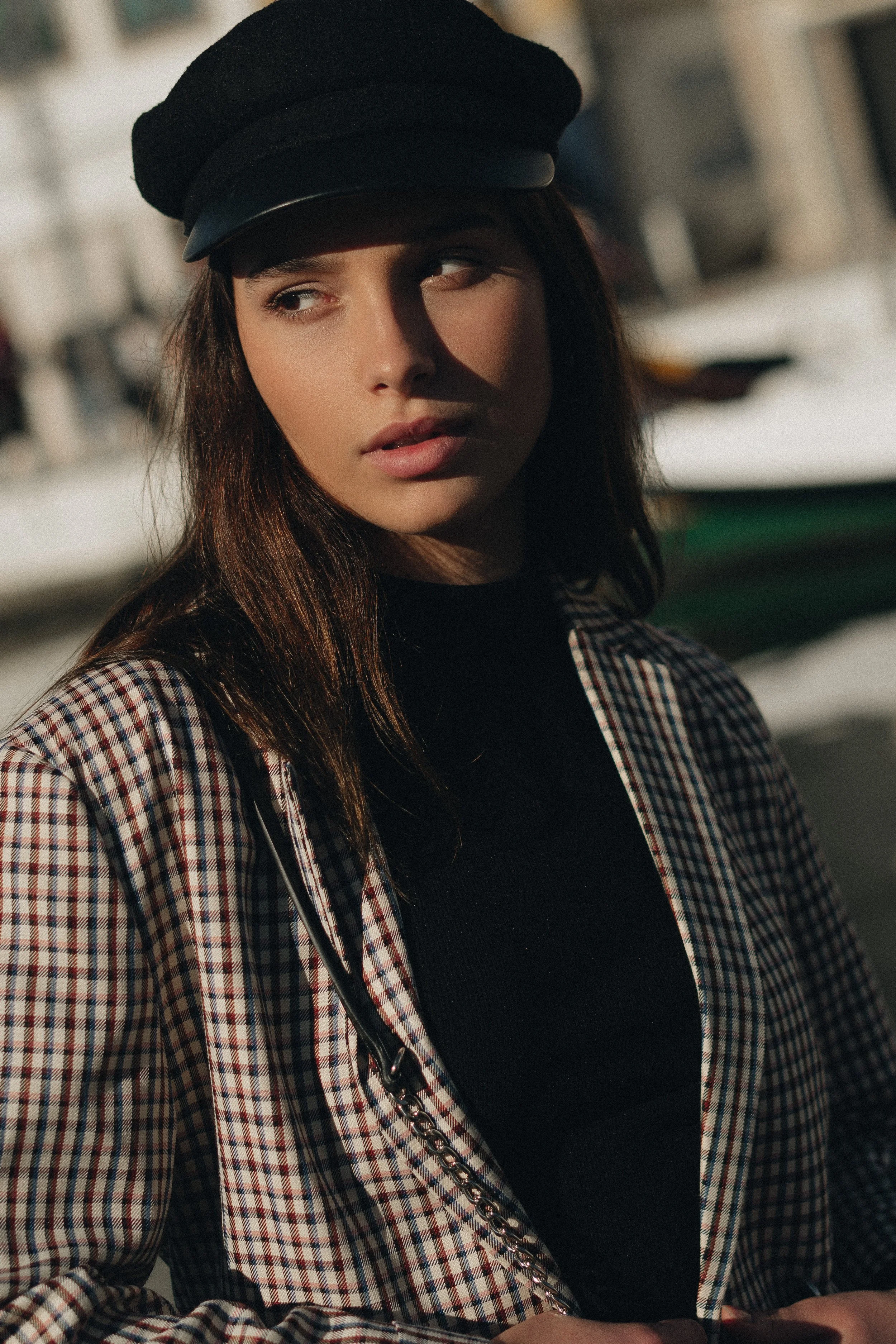 A young woman with brown hair wearing a black cap and a plaid jacket standing outdoors.