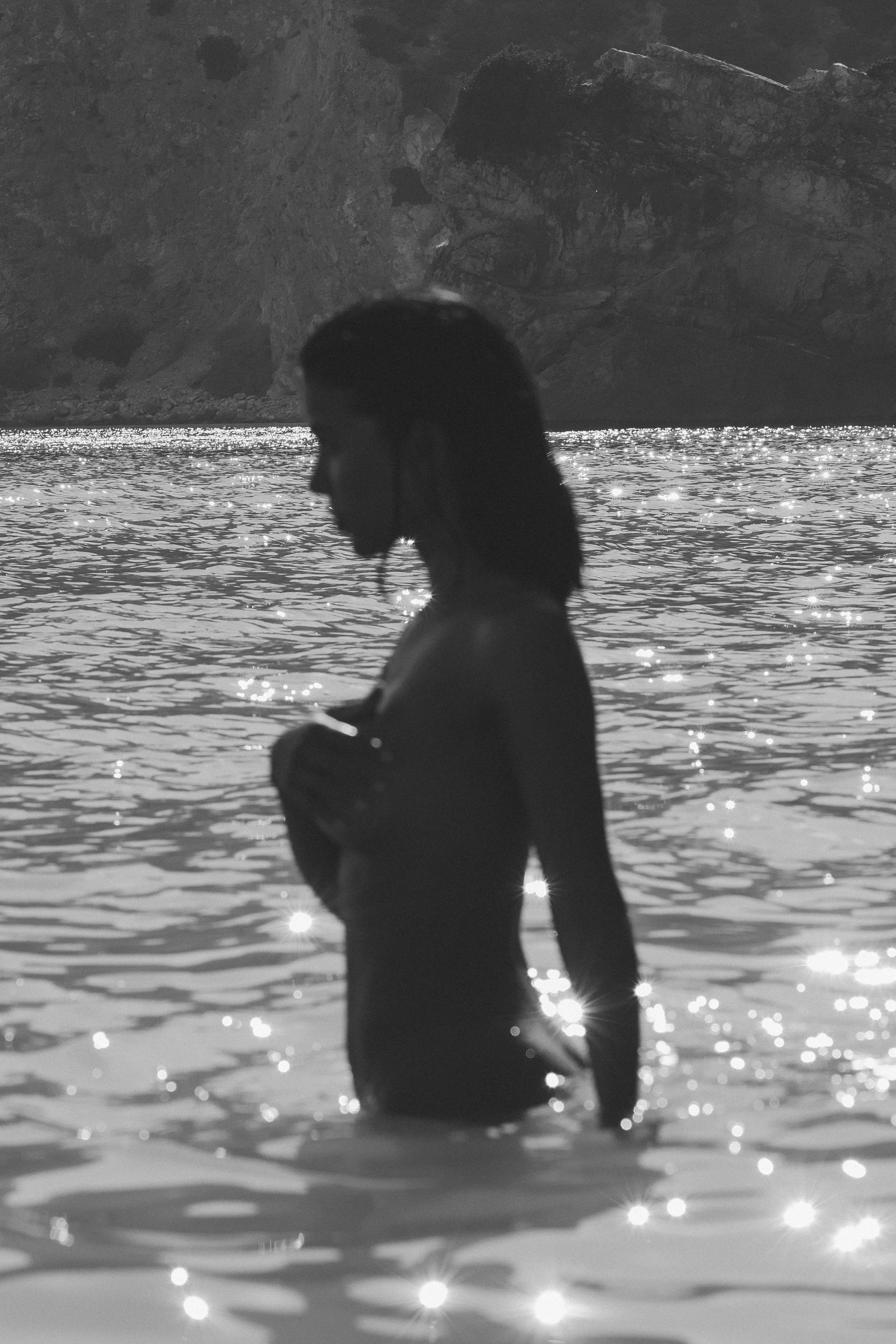 Silhouette of a woman standing in water with a mountain in the background. The sunlight reflects on the water's surface.