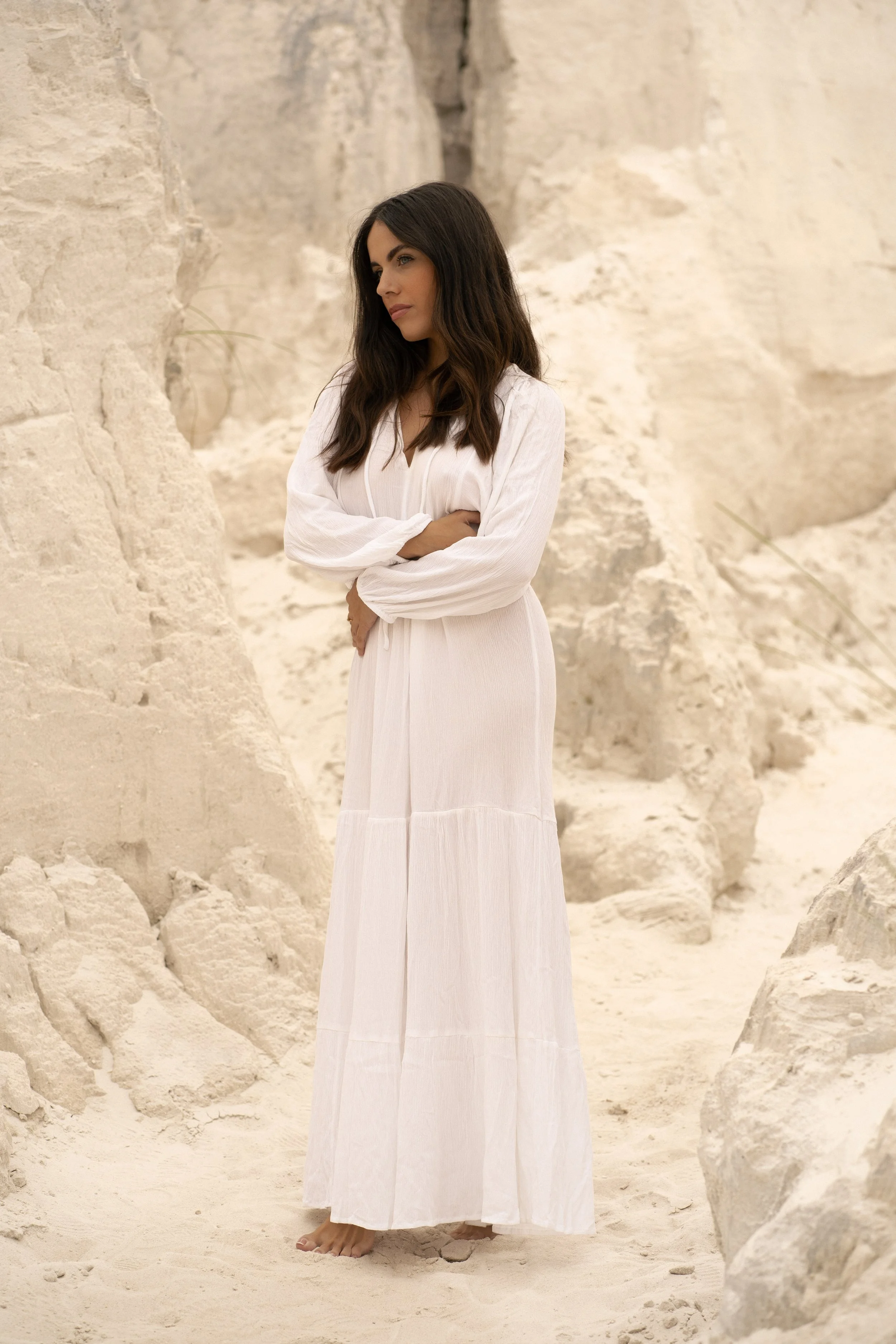 A woman with dark hair wearing a long white dress, standing barefoot in a sandy area with large rocks or cliffs in the background.