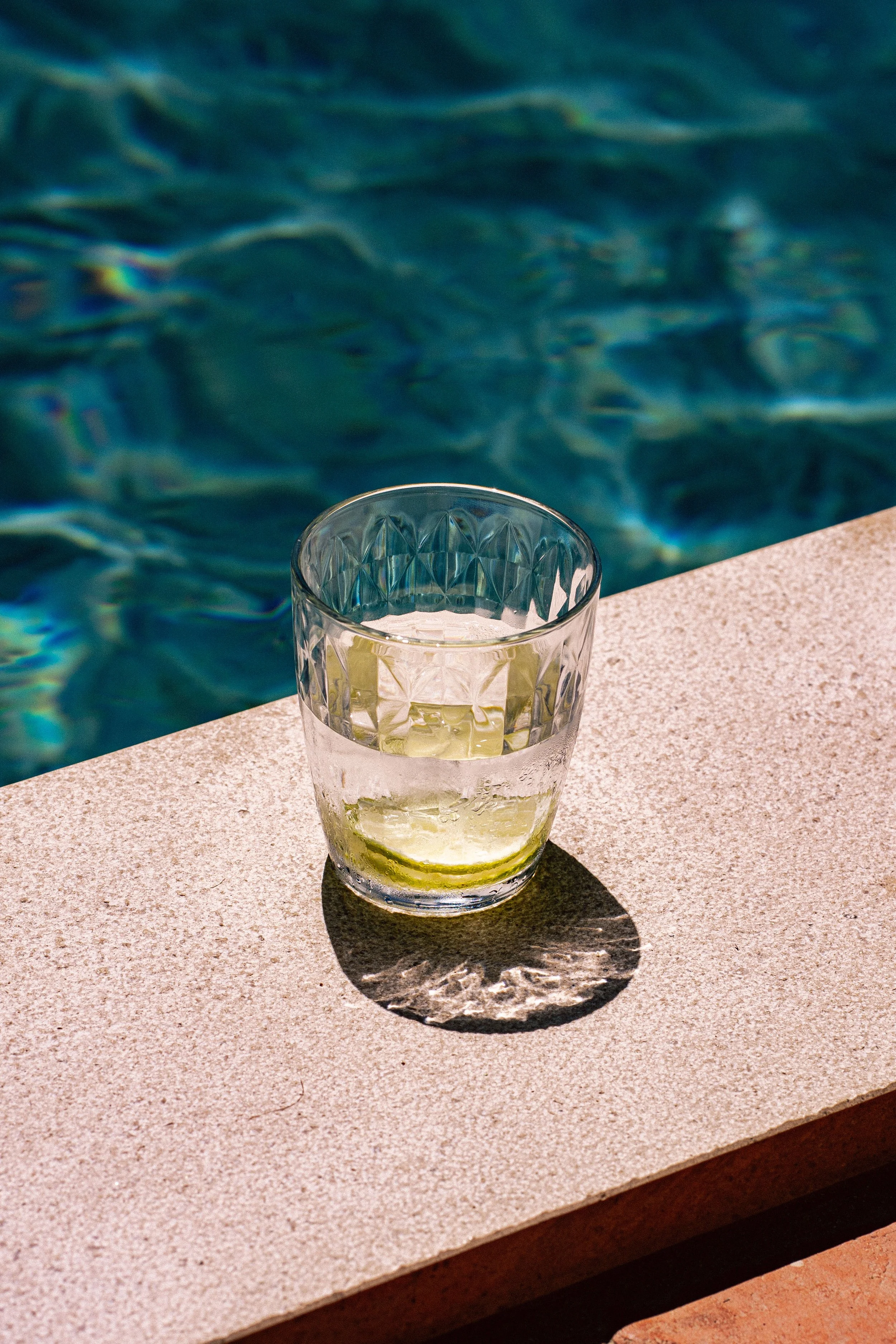 A glass of lemonade with a lemon wedge on the rim, placed on a beige outdoor table next to a swimming pool.