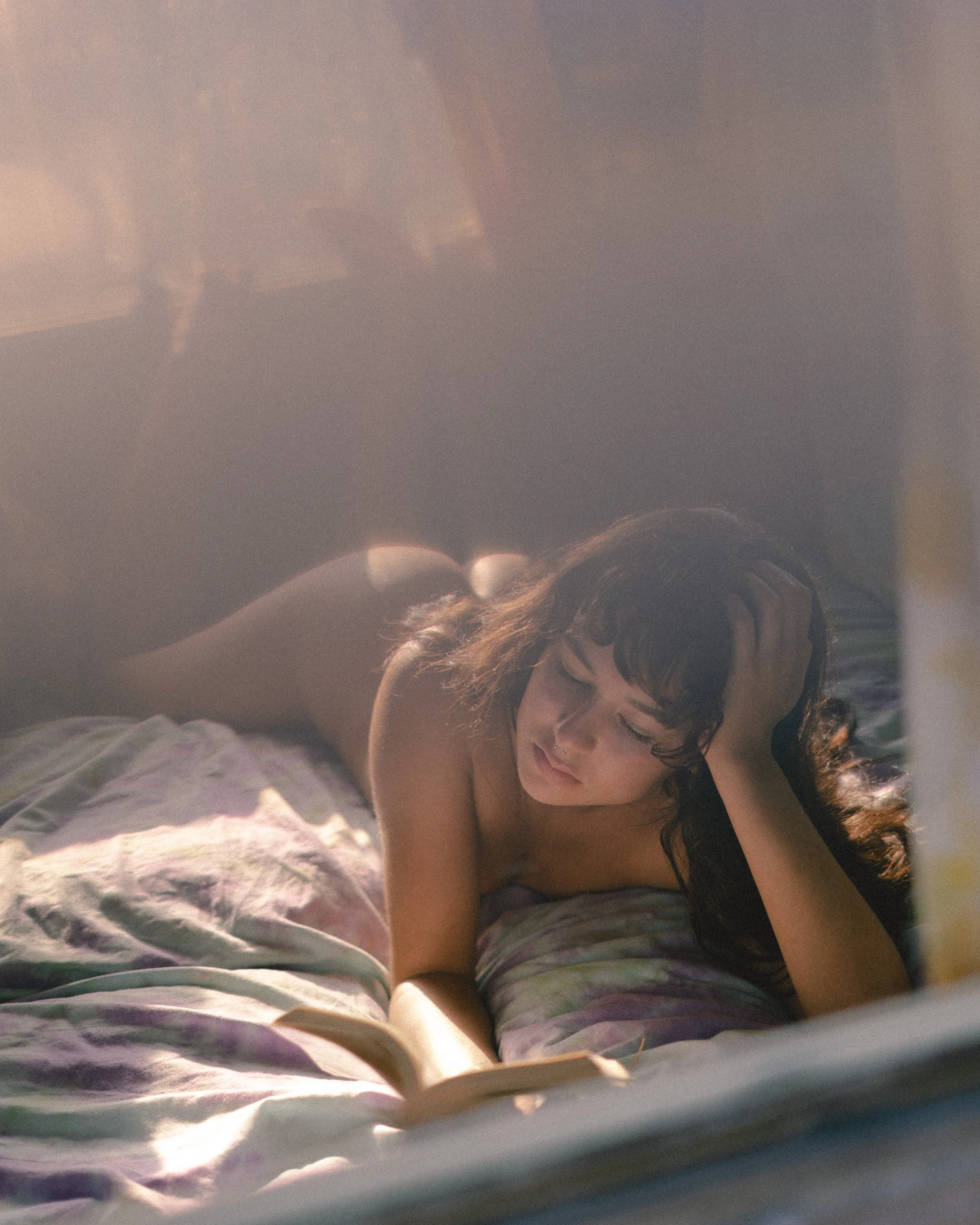 A woman with dark hair and a nose piercing is lying on her stomach on a bed, reading a book. Sunlight streams through a window, illuminating her face and the bed.