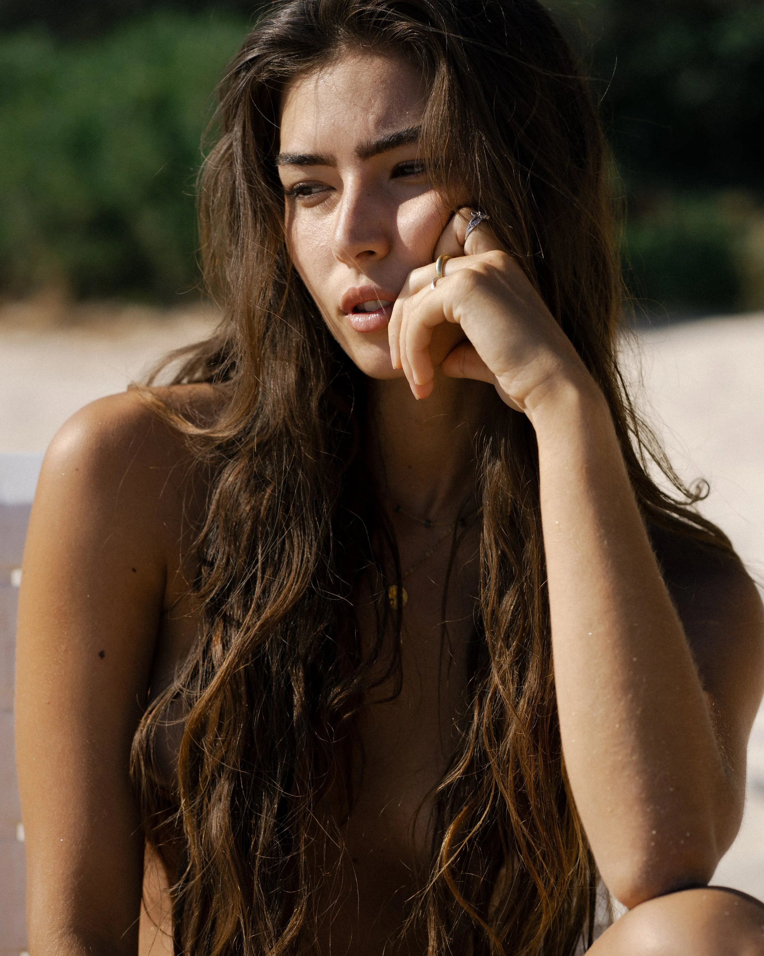 Close-up of a young woman with long brunette hair, wearing rings, with one hand near her face, outdoors on a sunny day, with blurred green background.
