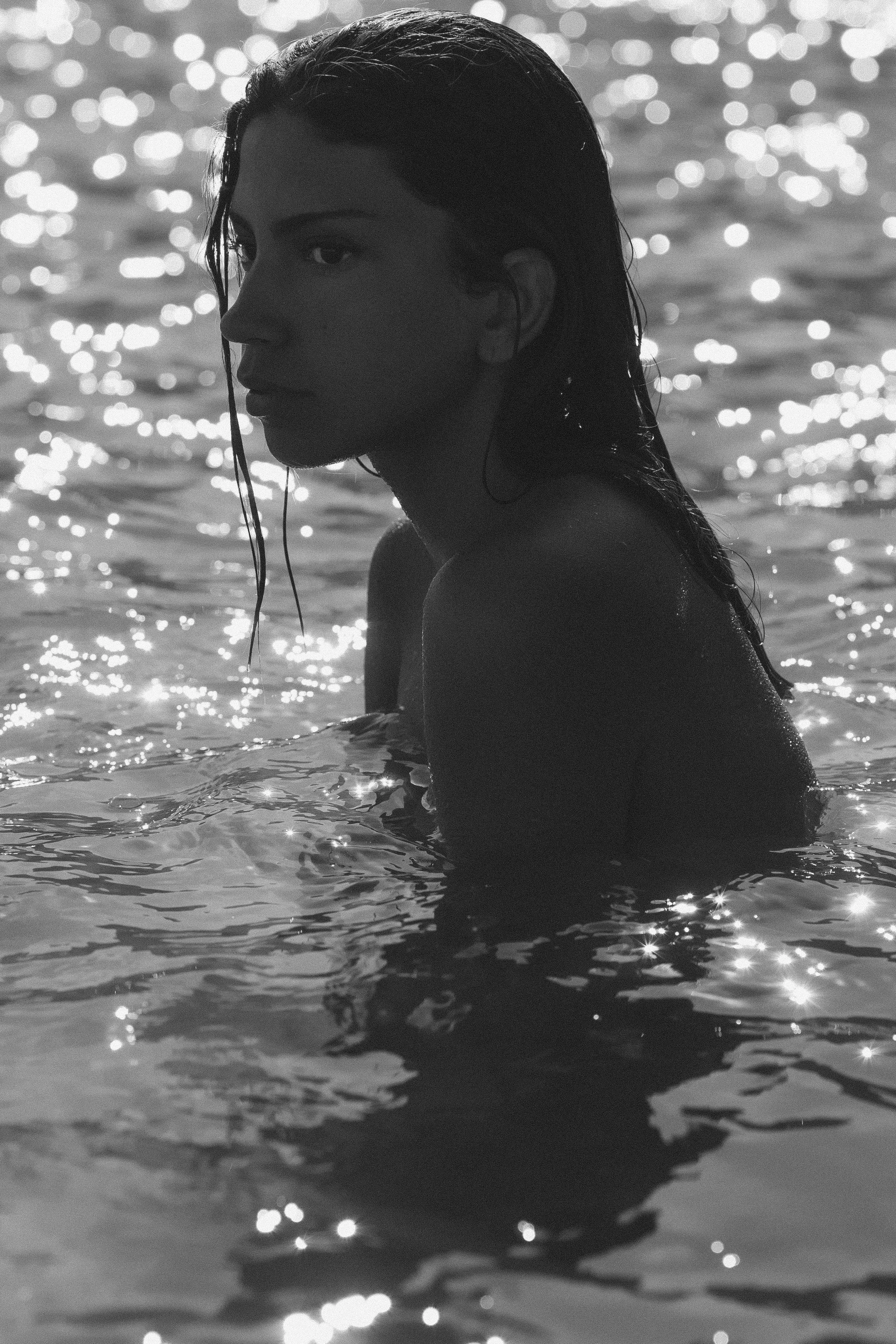 Black and white photo of a woman in water, with wet hair and a serious expression, backlit by sunlight, creating a shimmering bokeh effect in the background.