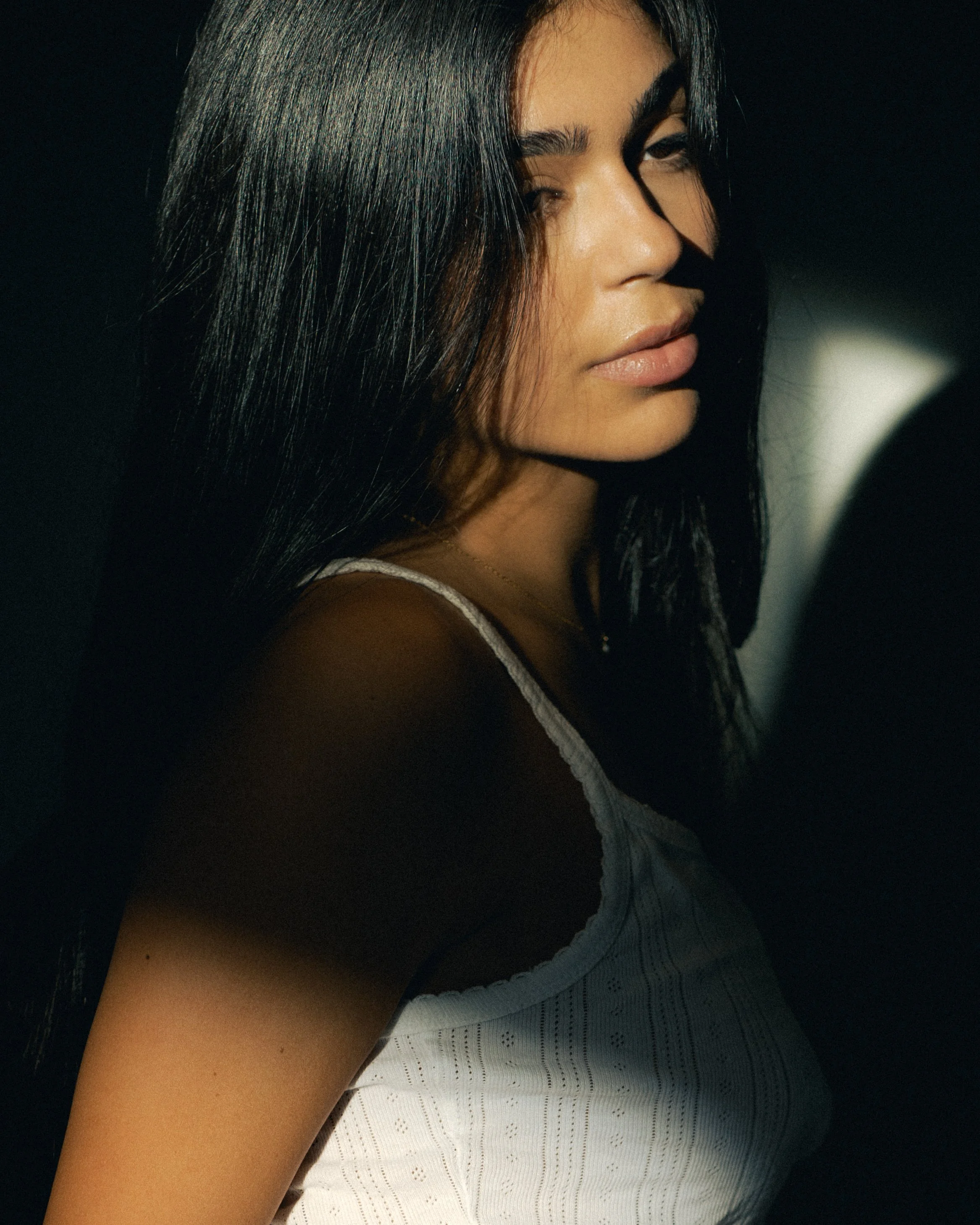 Close-up of a woman with dark hair, tan skin, and wearing a gray sleeveless top, standing in low lighting with a dark background.