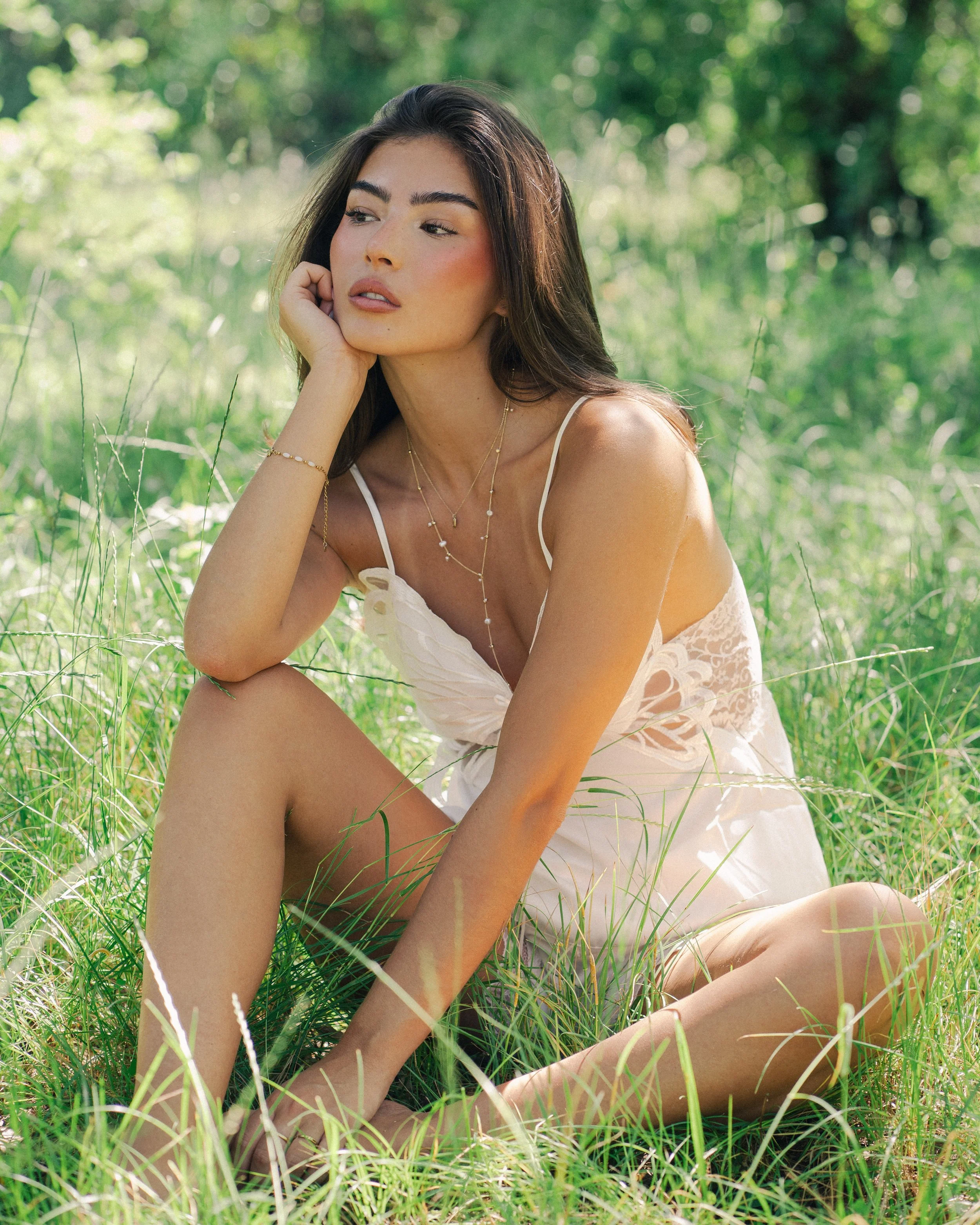 A woman with long dark hair sitting in tall green grass in a lush outdoor wooded area, resting her chin on her hand, wearing a white sleeveless dress and layered gold necklaces and bracelets.