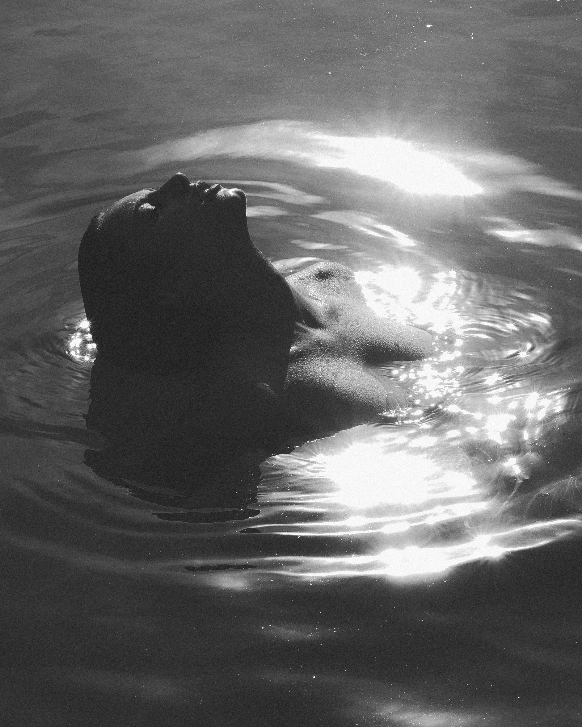 A person floating on their back in a body of water, with the sun reflecting off the surface, black and white photograph.