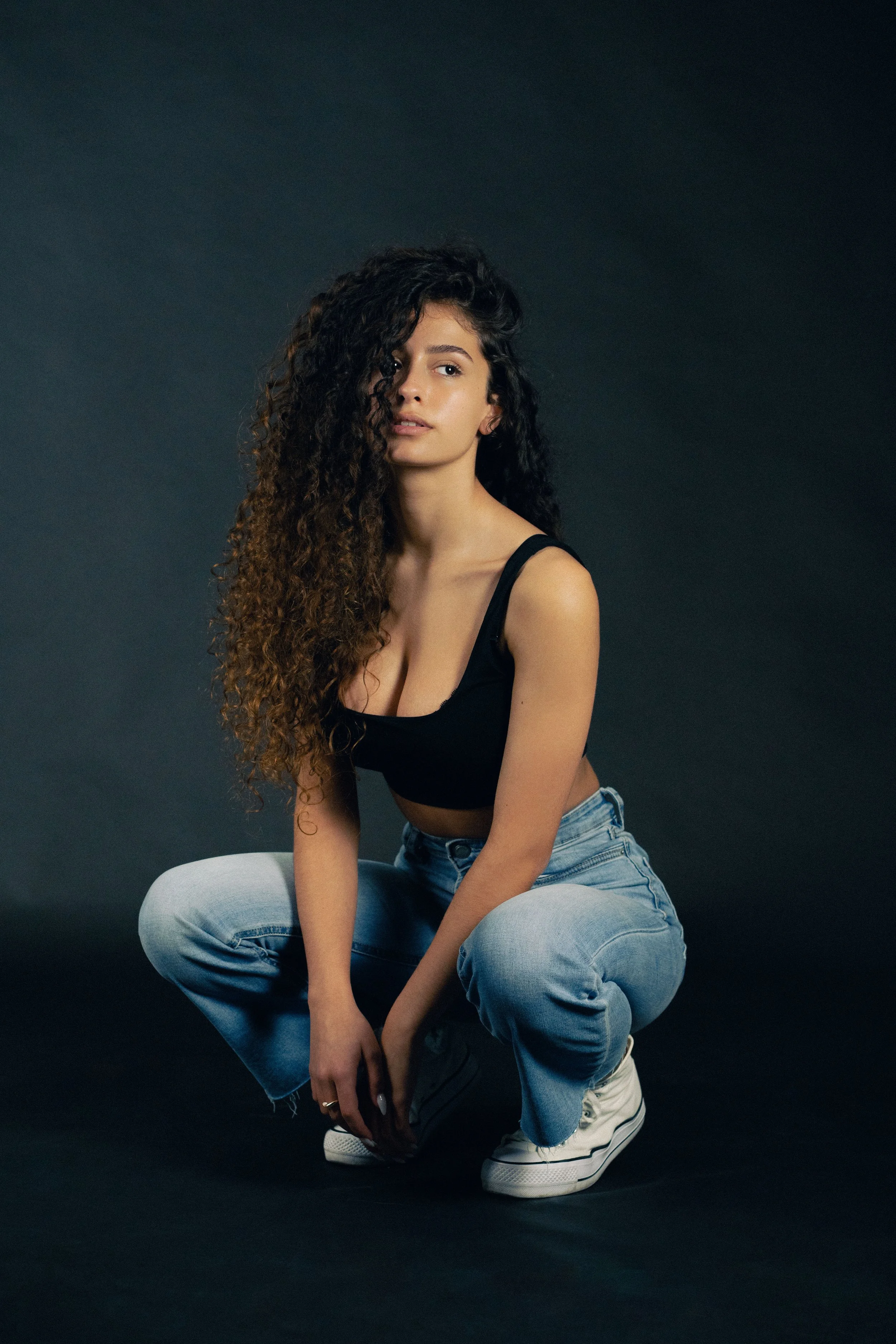 A young woman with long curly hair, wearing a black tank top, blue jeans, and white sneakers, squatting against a dark background.