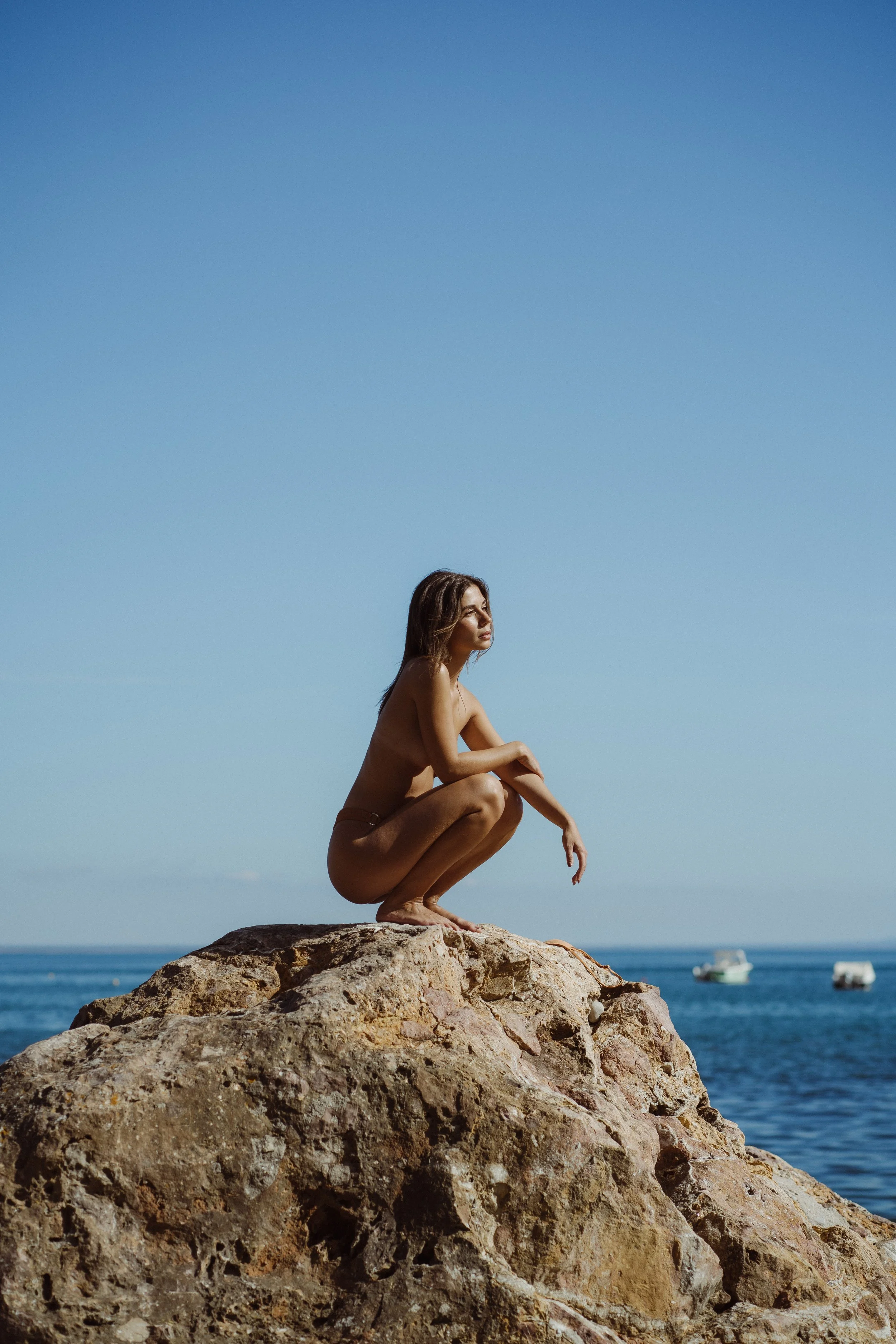 A woman crouching on a large rock at the beach, with an ocean and boats in the background, under a clear blue sky.